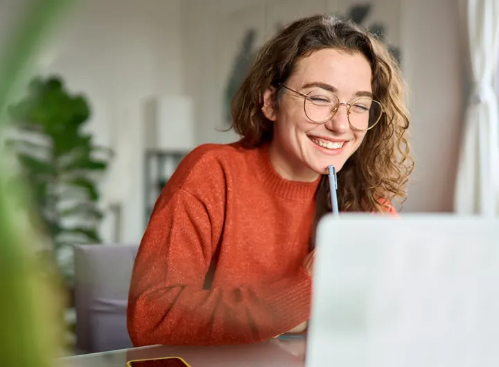 Person mit lockigem Haar und orangefarbenem Pullover sitzt an einem Tisch vor einem Laptop und trinkt durch einen Strohhalm.