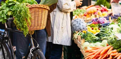 Fahrradkorb mit frischen Karotten, daneben Marktstand mit vielfältigem Gemüse und Obst, Menschen im Hintergrund.