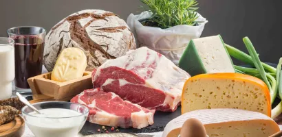 Verschiedene Käsesorten, rohes Fleisch, ein Laib Brot, ein Glas Rotwein, ein Ei und frische Kräuter auf einem Tisch arrangiert.