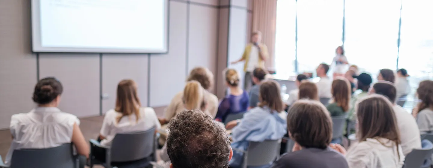Gruppe von Menschen sitzt in einem Seminarraum und blickt auf eine Projektionsfläche, vorne steht eine Person und hält einen Vortrag.