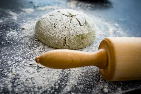Runder, mit Mehl bestäubter Brotteig auf dunkler Arbeitsfläche neben einem Holz-Nudelholz.