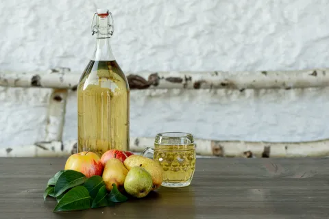Große Glasflasche mit hellem Most, Glas mit Most, mehrere Birnen und Äpfel mit grünen Blättern auf dunklem Holztisch vor weißer Wand.