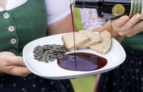 Dunkelbraunes Öl wird auf einen weißen Teller mit Kürbiskernen und Brotscheiben gegossen, zwei Personen in Trachten halten den Teller und die Flasche.