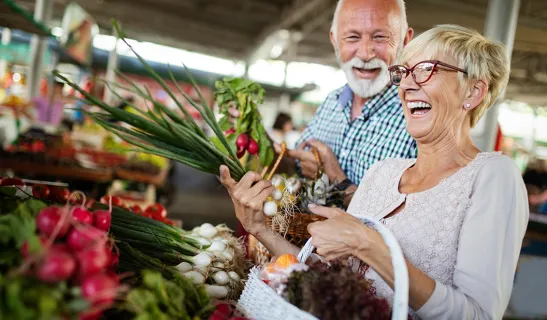Zwei Personen beim Einkauf auf einem Markt, eine hält Frühlingszwiebeln, die andere einen Korb mit Gemüse.