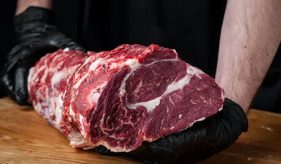 Zwei Hände mit schwarzen Handschuhen halten ein großes, marmoriertes rotes Fleischstück auf einem Holzbrett.