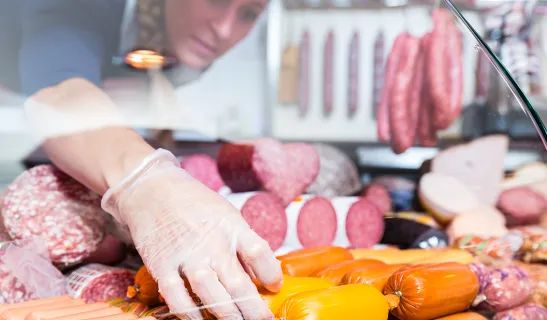 Hand mit Einweghandschuh greift in einer Fleischtheke nach orangefarbenen Würstchen, im Hintergrund verschiedene Wurstsorten.