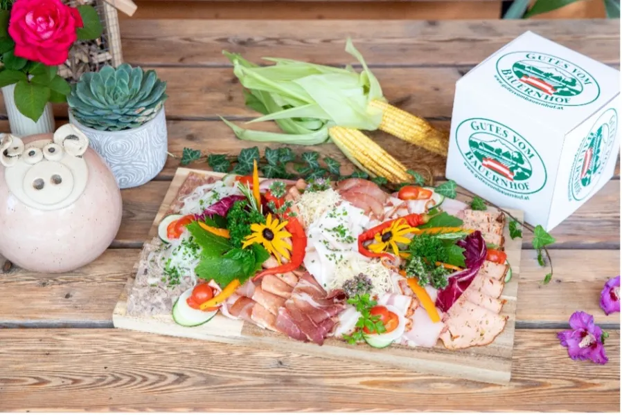 Brett mit verschiedenen Aufschnittsorten, Kräutern und Gemüsestücken, daneben Maiskolben und ein weißer Würfel mit dem Schriftzug 'GUTES VOM BAUERNHOF'.