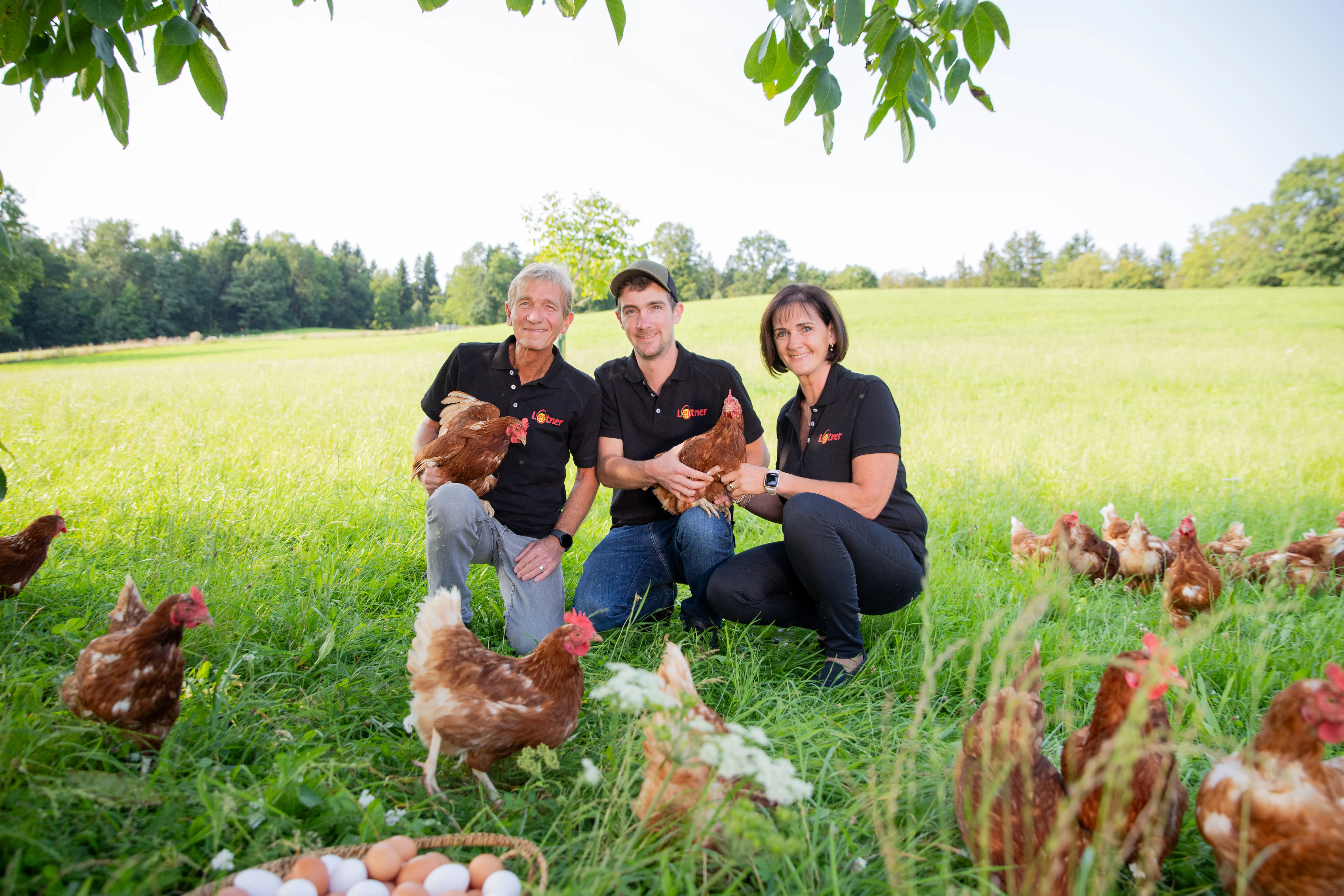 Ein Mann und eine Frau mit ihrem erwachsenen Sohn hocken auf einer grünen Wiese mit verschieden farbigen Hennen, sie halten zwei Hühner im Arm