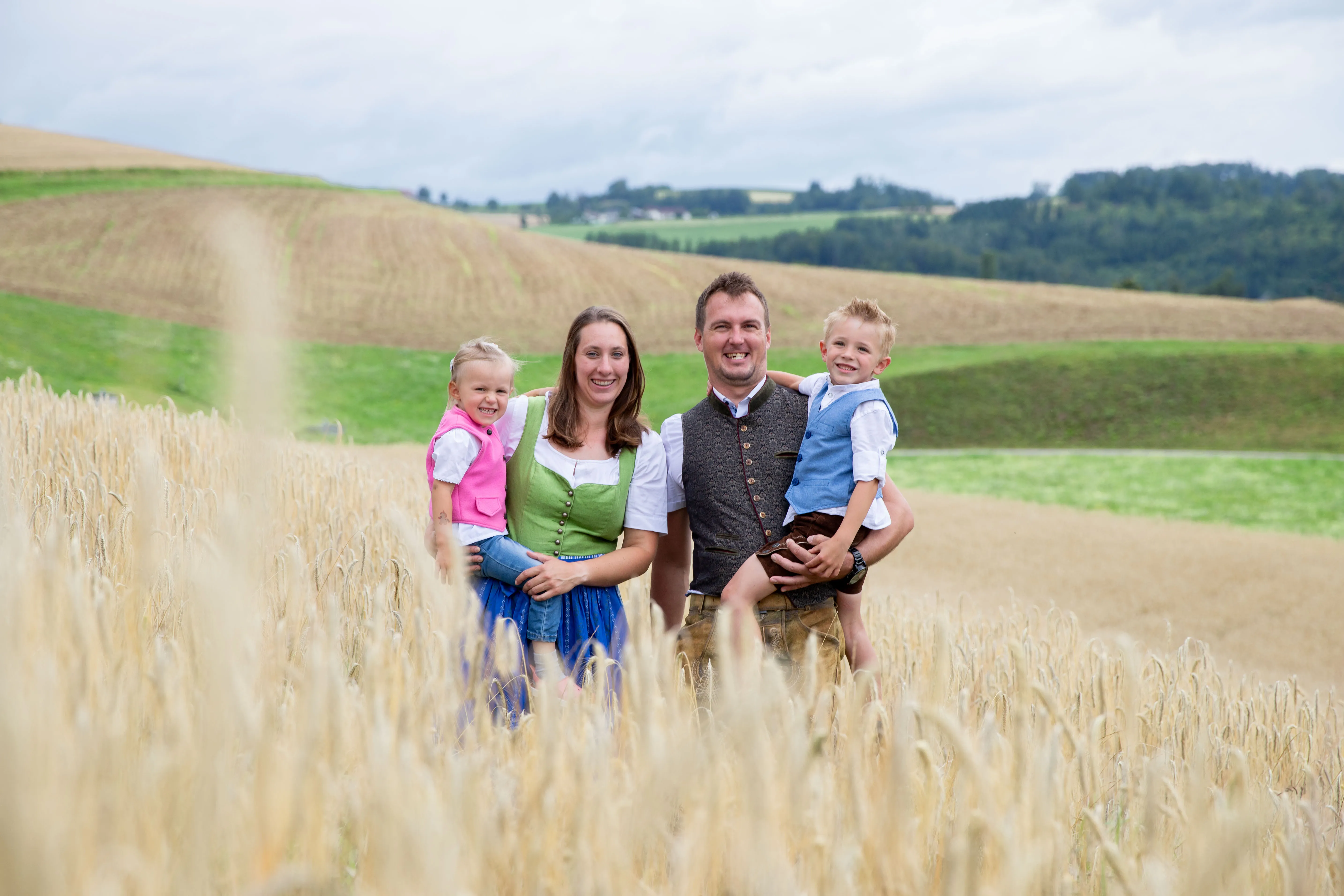 Ein Mann und eine Frau mit zwei Kindern auf dem Arm stehen in einem Feld, im Hintergrund grüne Wiese, Wald und blauer Himmel