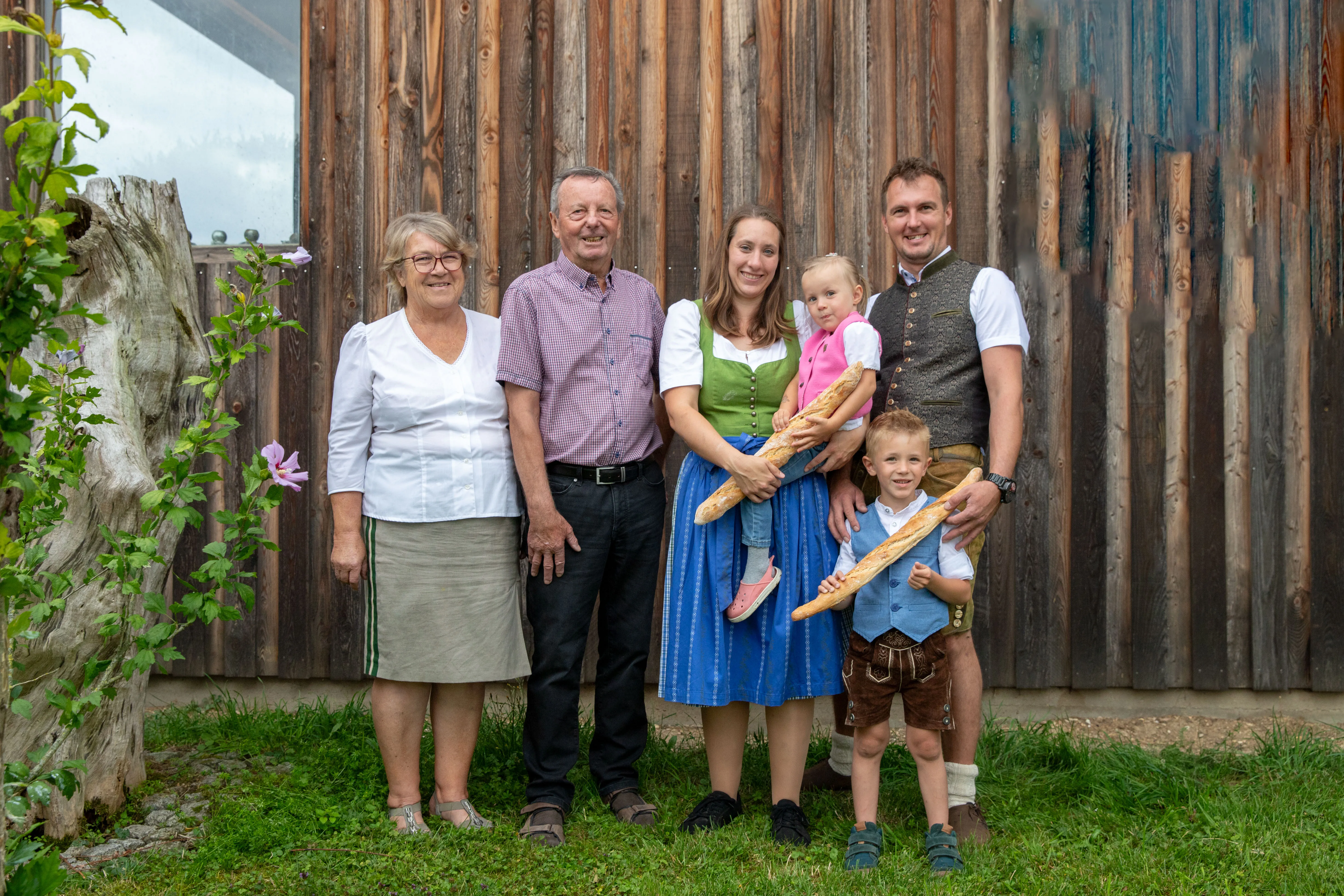 Vier Erwachsene mit Kindern stehen vor einem braunen Holztor, die Kinder halten Weißbrot in den Händen