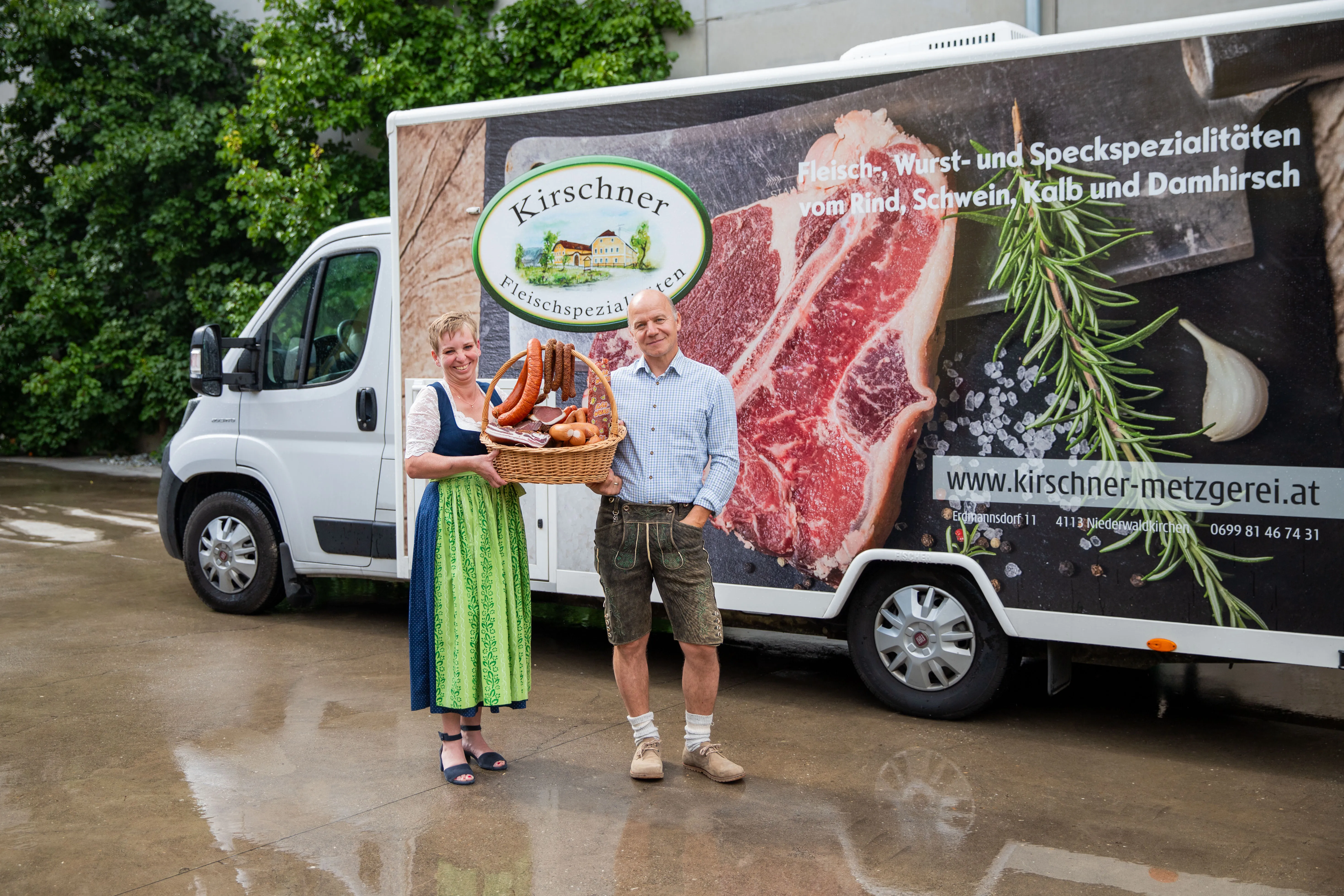 Ein Mann und eine Frau stehen im Vordergrund eines großen Lieferwagen mit Aufschrift, sie halten einen Korb mit Fleischprodukten im Arm