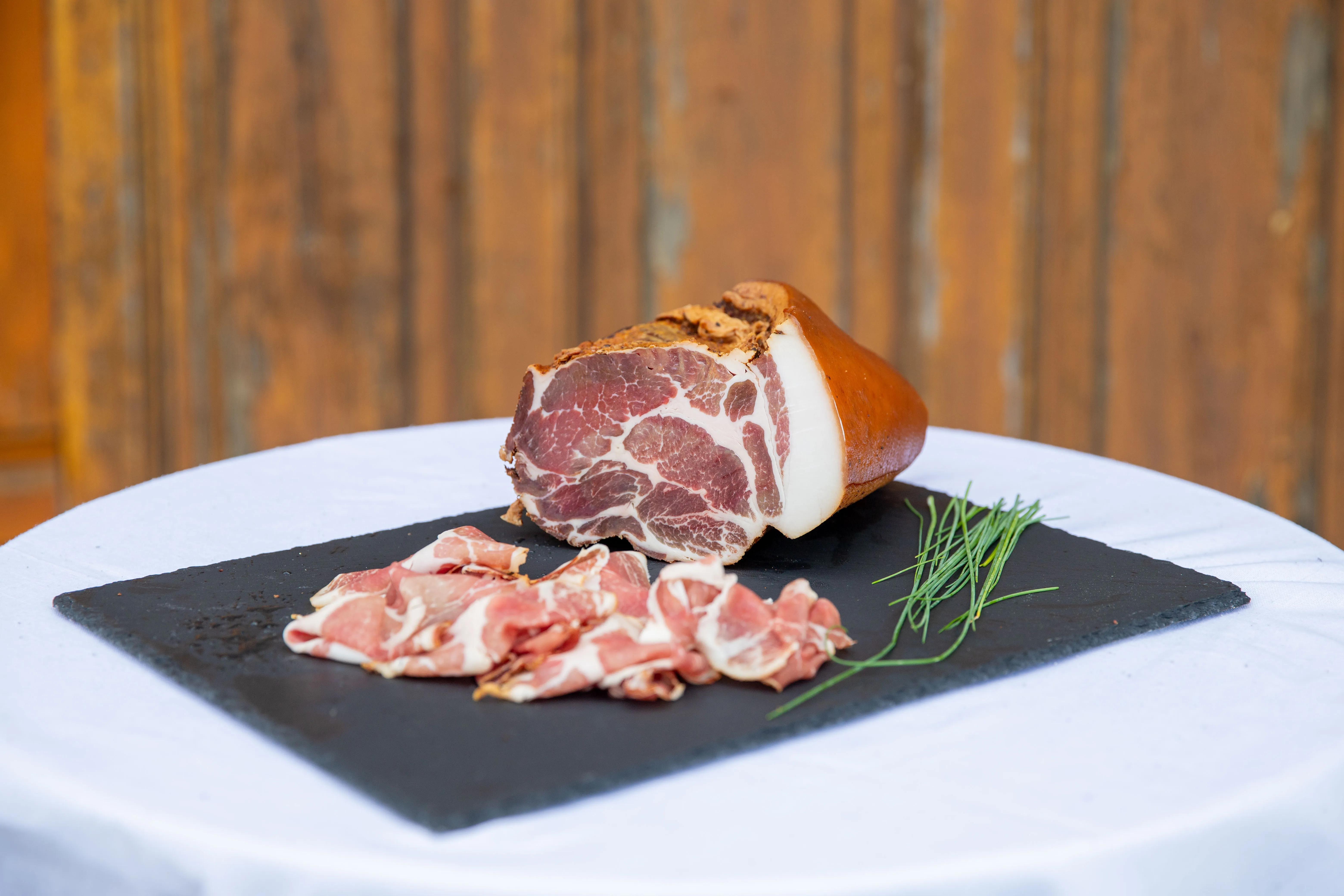 Aufgeschnittener Speck auf einer schwarzen Platte, weißes Tischtuch und braune Wand im Hintergrund