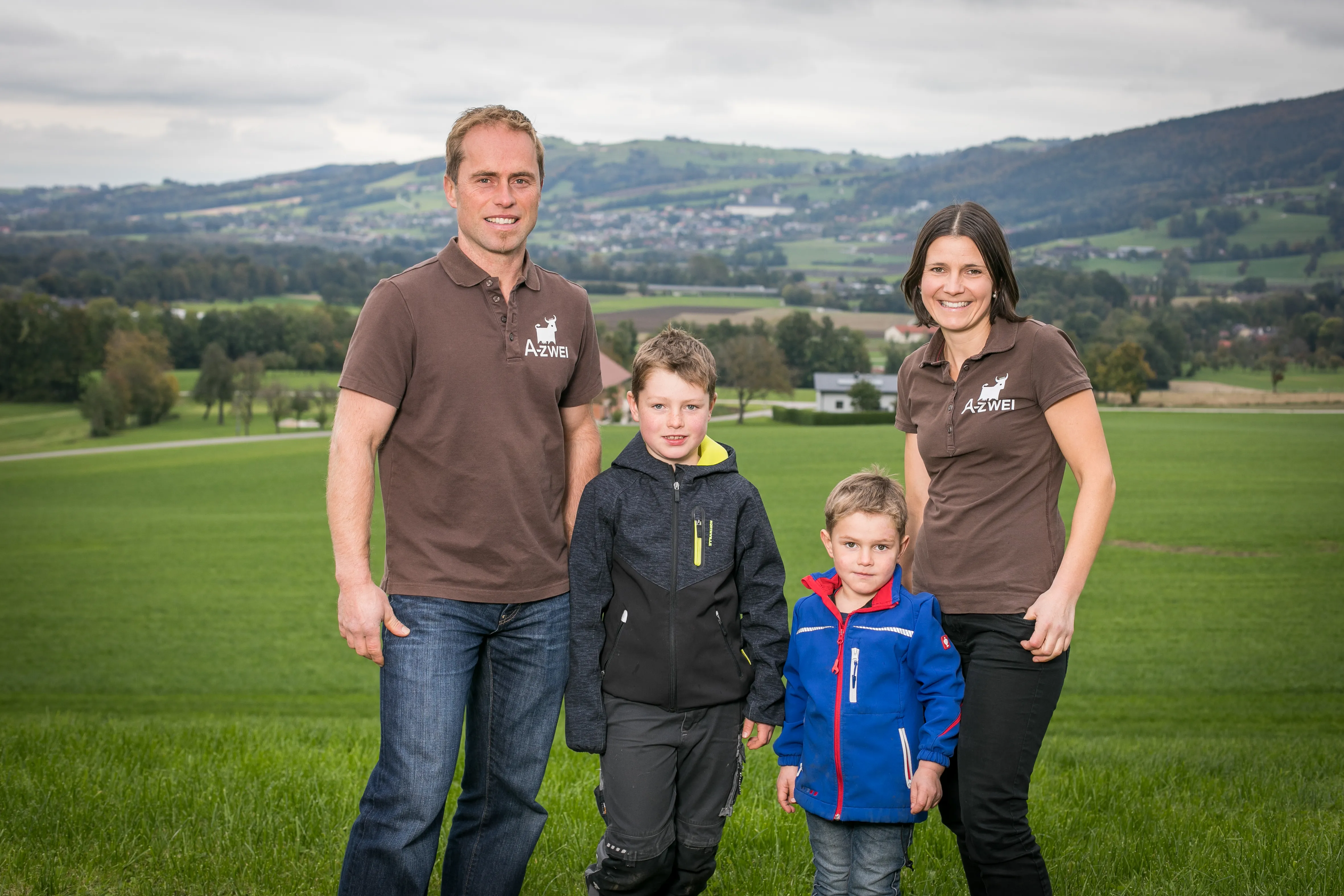 Ein Mann, eine Frau und zwei Kinder stehen auf einer Wiese, im Hintergrund Hügel, Häuser und Bäume