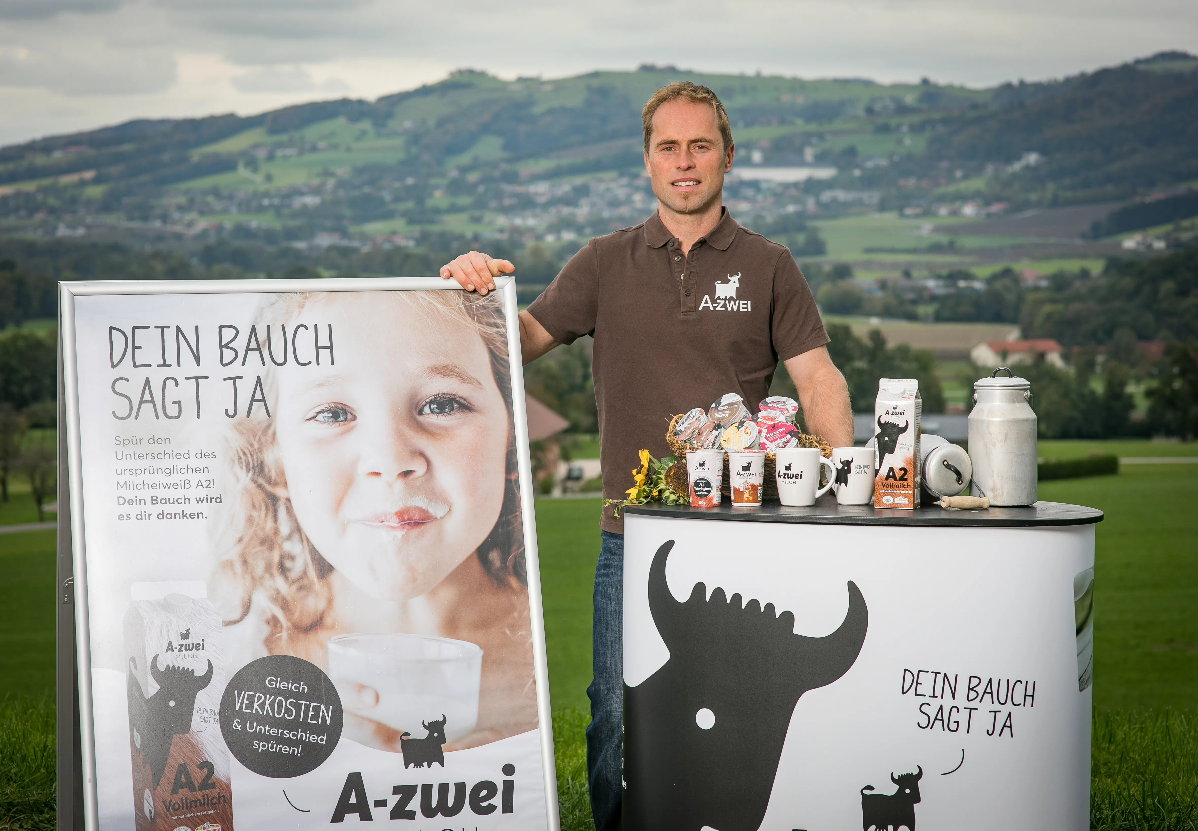 Ein Mann steht auf einer Wiese, im Hintergrund Hügel und Bäume, im Vordergrund eine Tafel mit einem Mädchen drauf und ein Stehtisch mit diversen Produkten