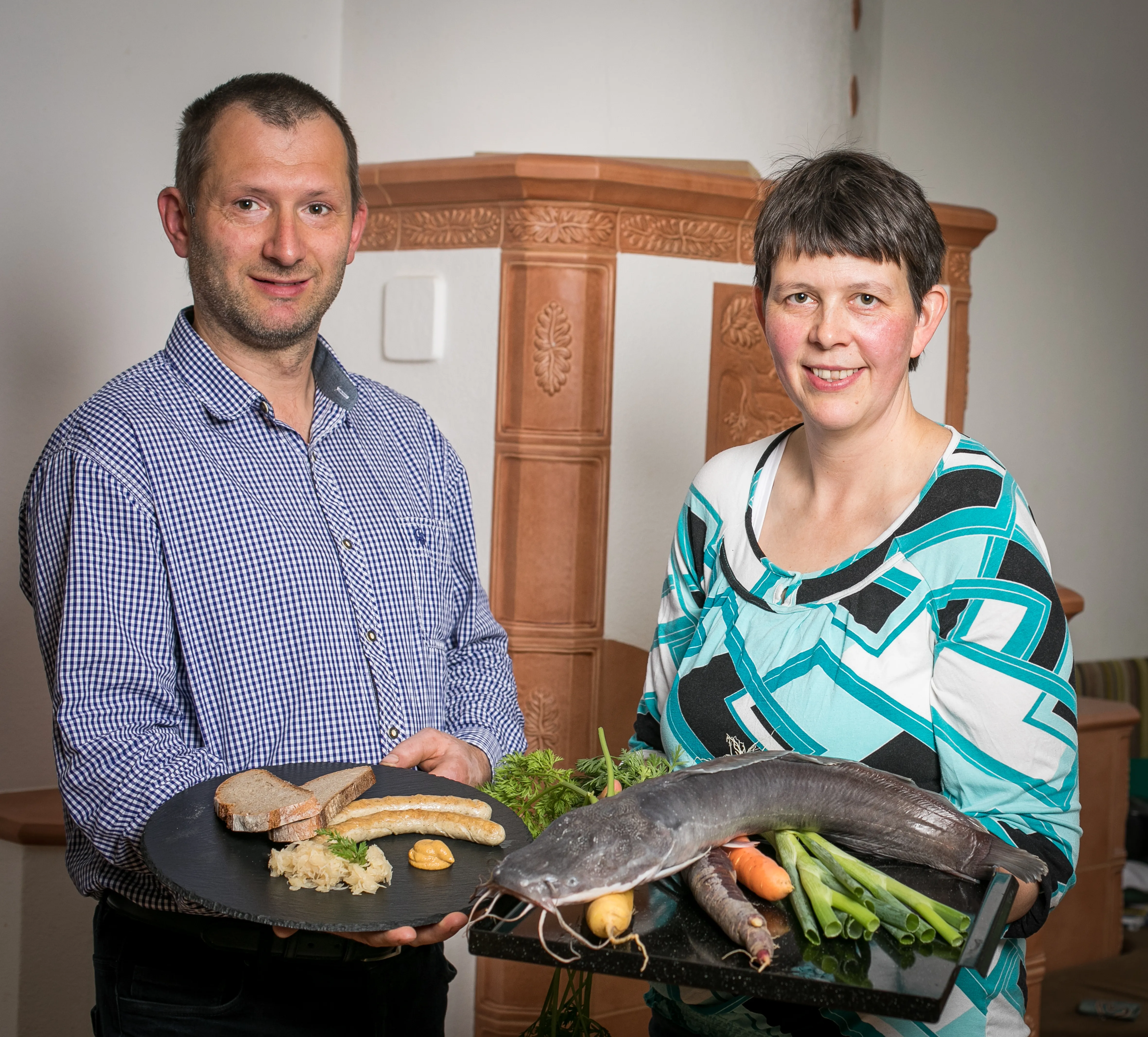 Ein Mann im blauen Hemd und eine Frau in bunter Bluse stehen vor einem Kachelofen und halten jeweils eine Platte mit einem Fisch und Produkten in den Händen