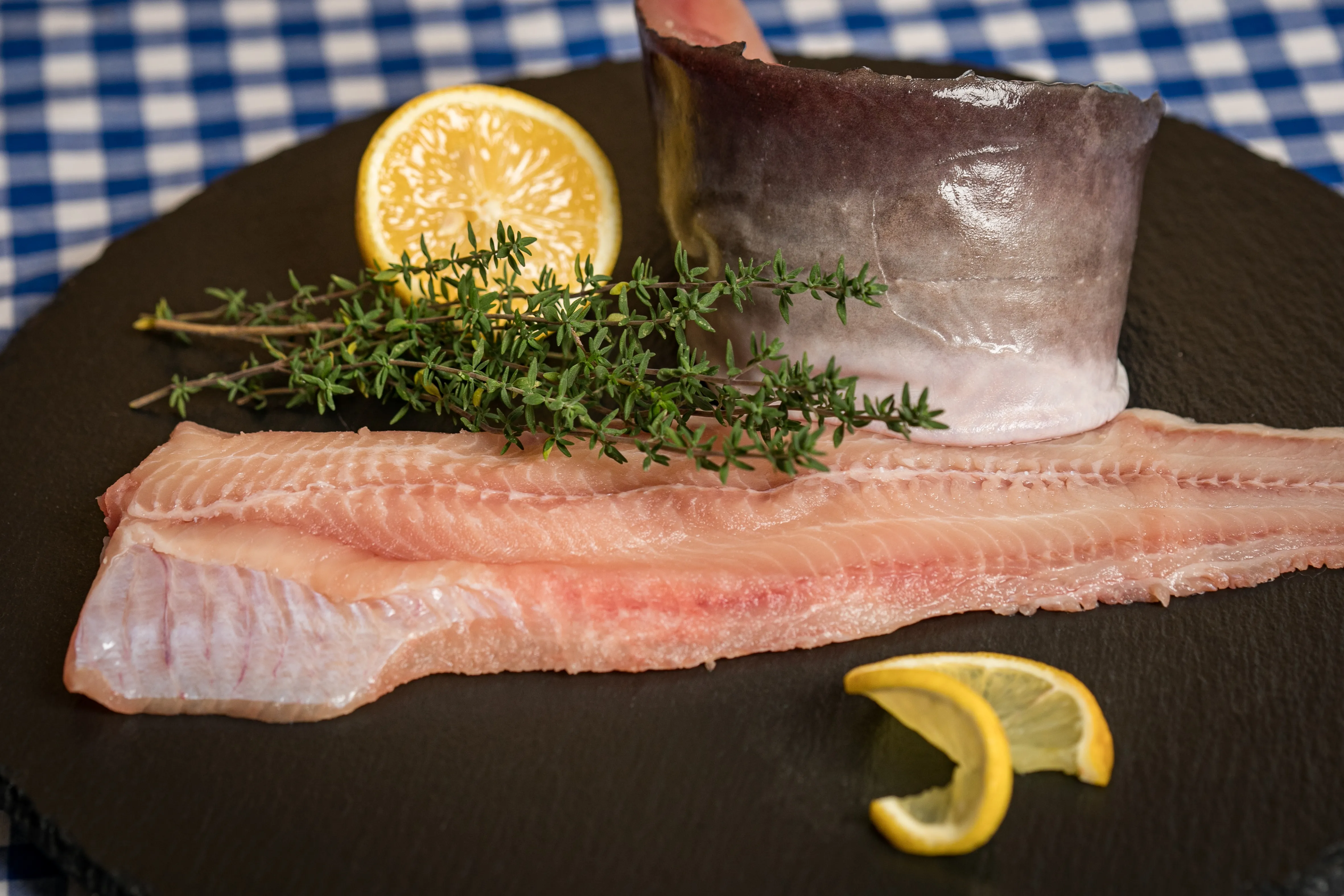 Auf einer schwarzen Platte liegt ein Fischfilet und ein gerolltes Filet, garniert mit einer Zitrone und Kräuter