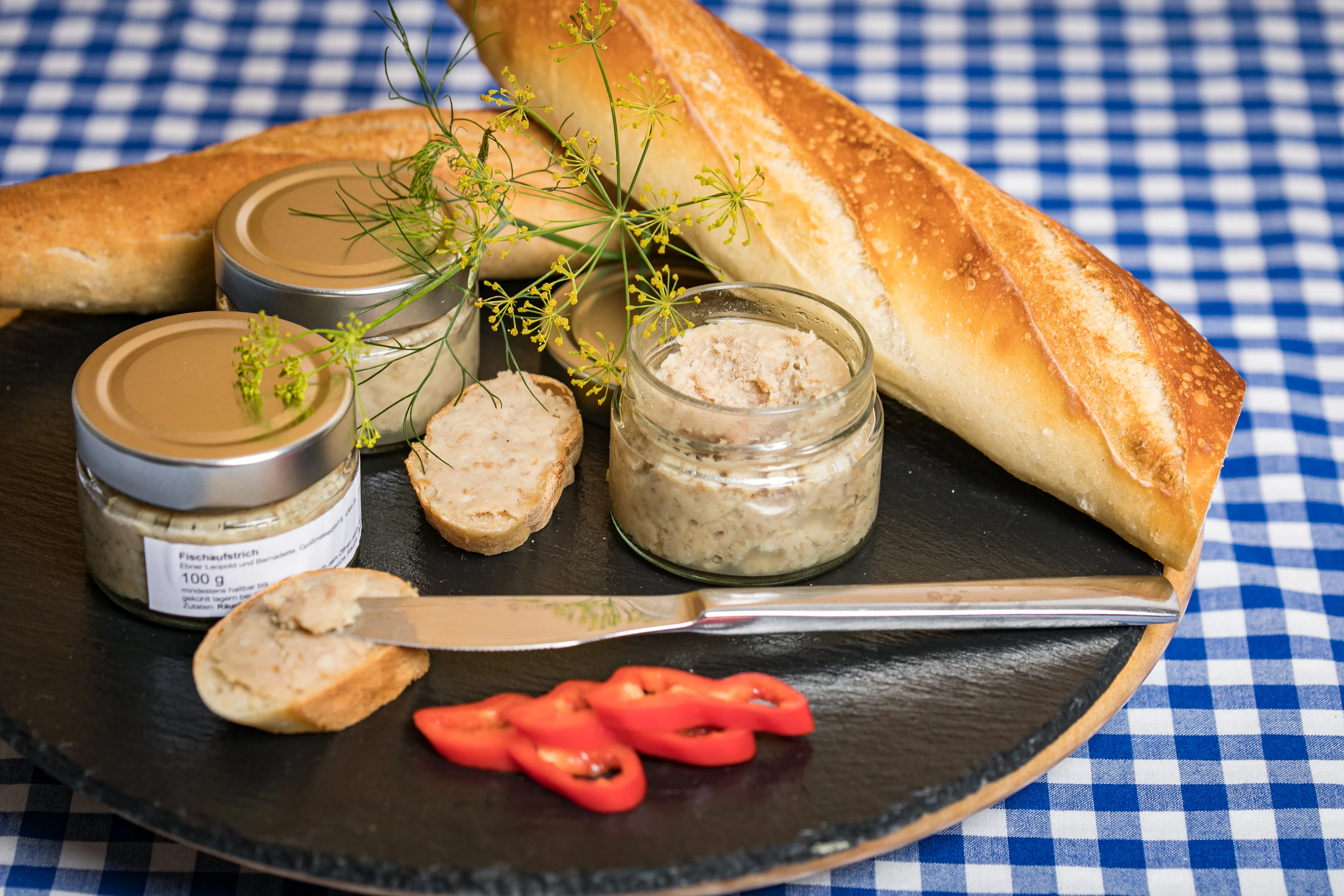 Auf einer blau/weiß karierten Tischdecke steht ein dunkler Teller mit drei Gläsern mit Fischaufstrich, ein Baguette liegt auf dem Teller, zudem ein Messer und roter Paprika und Kräuter