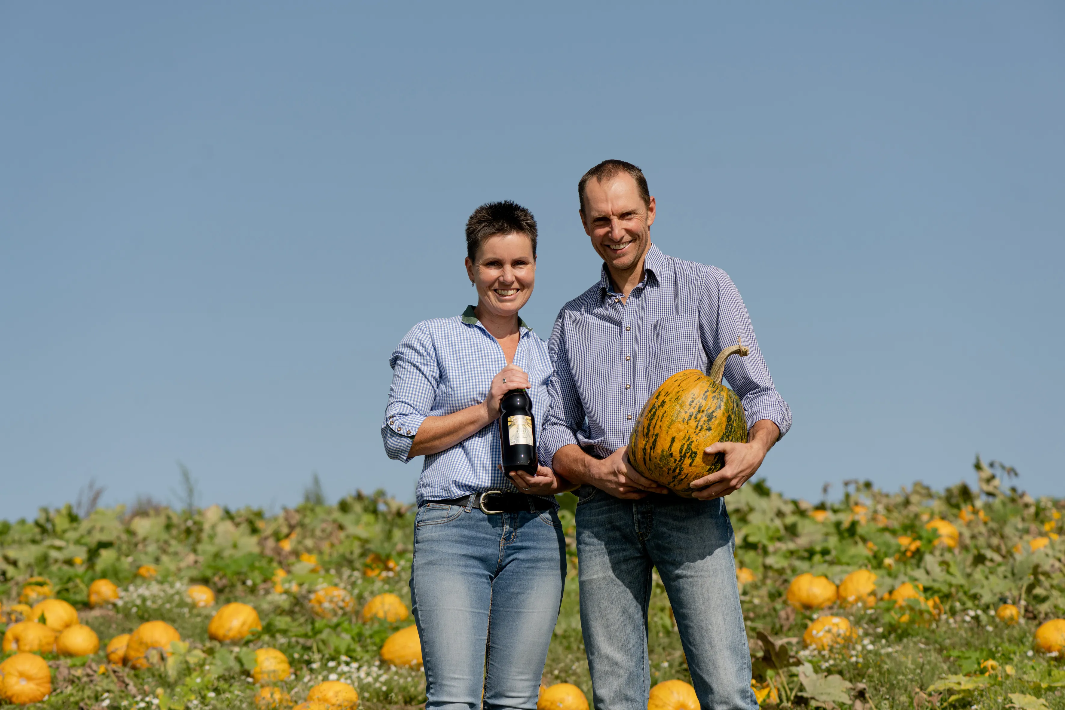 Ein Man und eine Frau stehen vor einem Kürbisfeld, viele orange Kürbisse im Hintergrund, der Mann hält einen Kürbis in den Händen, die Frau eine Flasche