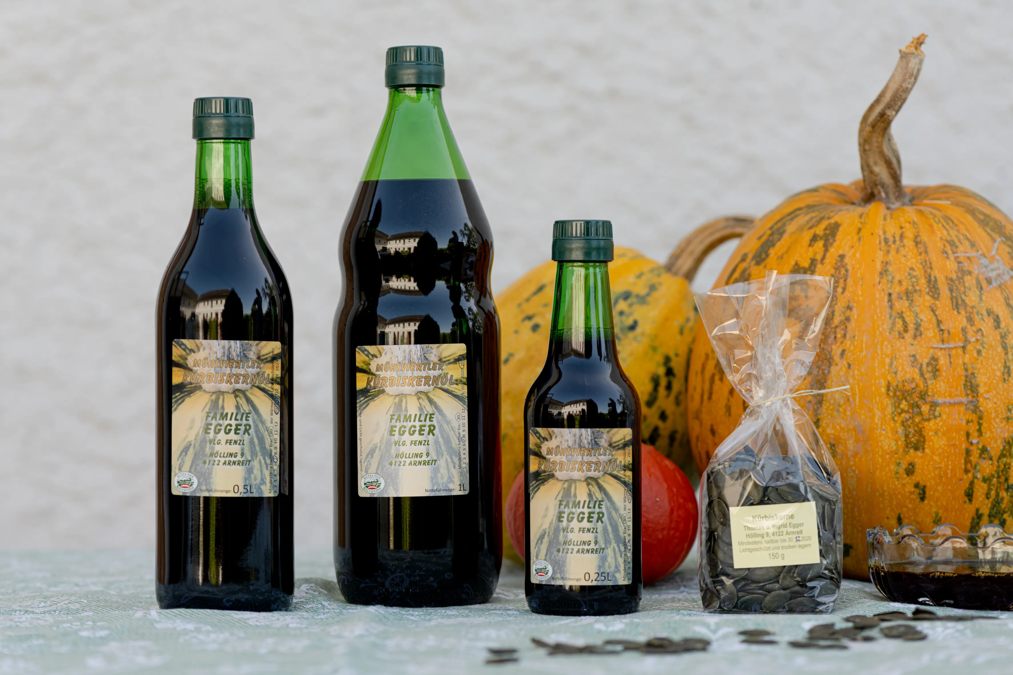 Drei verschieden große Flaschen, zwei Kürbisse und ein Sackerl mit Kernen stehen auf einem grauen Untergrund, grauer Hintergrund