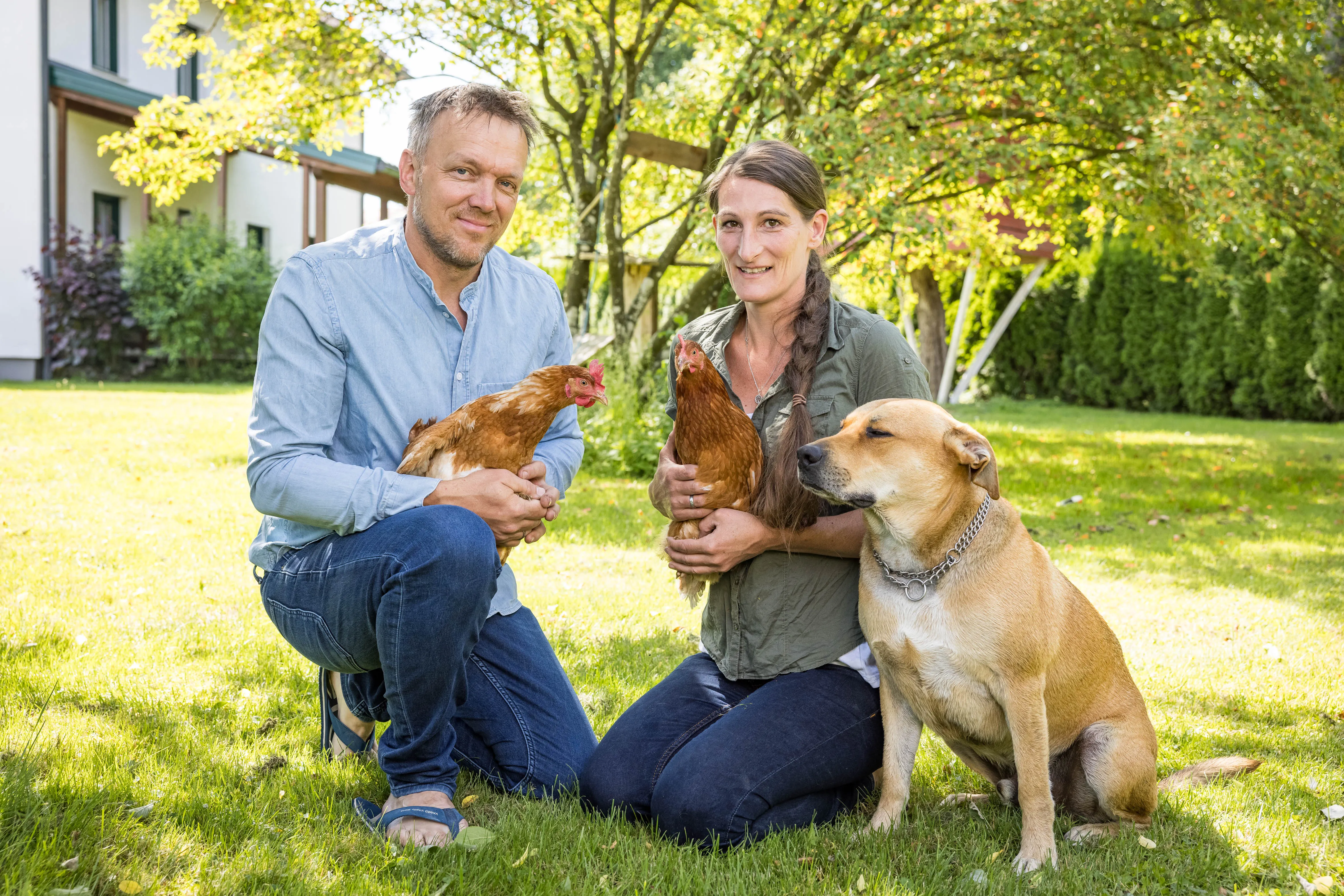 Ein Mann in blauem Hemd und Jeans und eine Frau mit grüner Bluse und Jeans knien in der Wiese und halten jeweils ein Huhn in der Hand. Daneben sitzt ein brauner Hund. Dahinter grüne Bäume und ein weißes Wohngebäude.