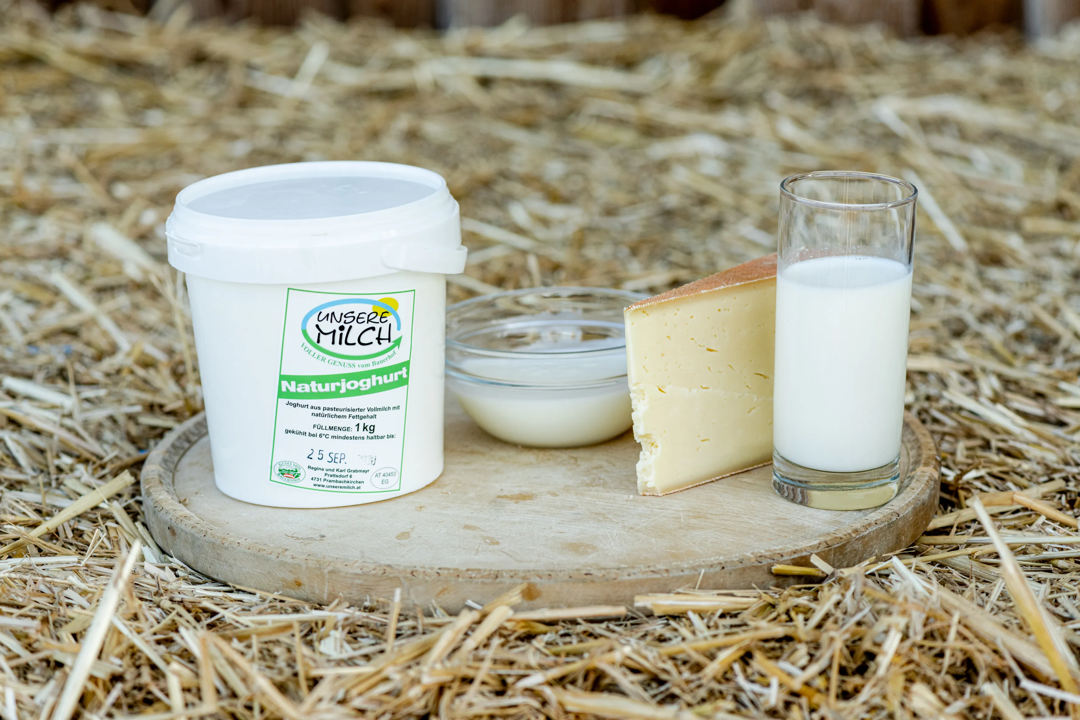 Ein weißer Joghurtbecher, eine Schale, ein Stück Käse und ein Glas Milch stehen auf einem Holzbrett, welches im Stroh steht