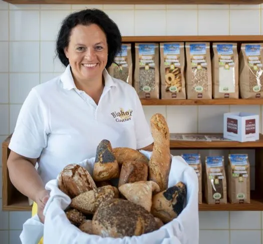 Eine Frau mit schwarzen Haaren und weißer Kleidung hält einen Brotkorb mit weißem Tuch und verschiedenen Brotsorten in der Hand und stehen vor einem Holzregal mit verschiedenen Verpackungen. Dahinter Wand mit weißen Fliesen.