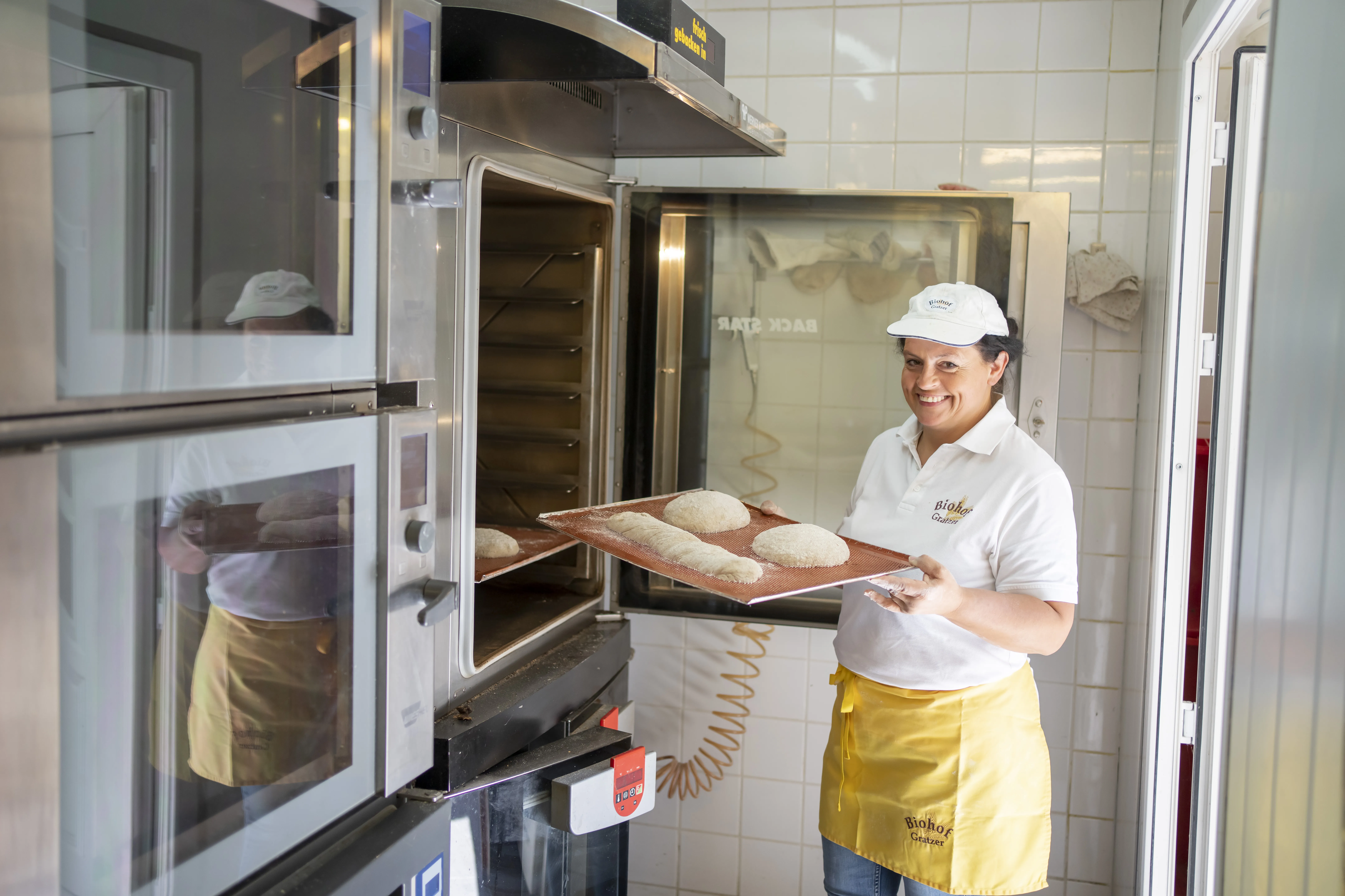 Eine Frau mit weißem Kapperl und weißer Kleidung, gelbe Schürze steht mit einem Backblech mit Brot vor einem großen Backofen, welcher geöffnet ist. Daneben weitere Backöfen.