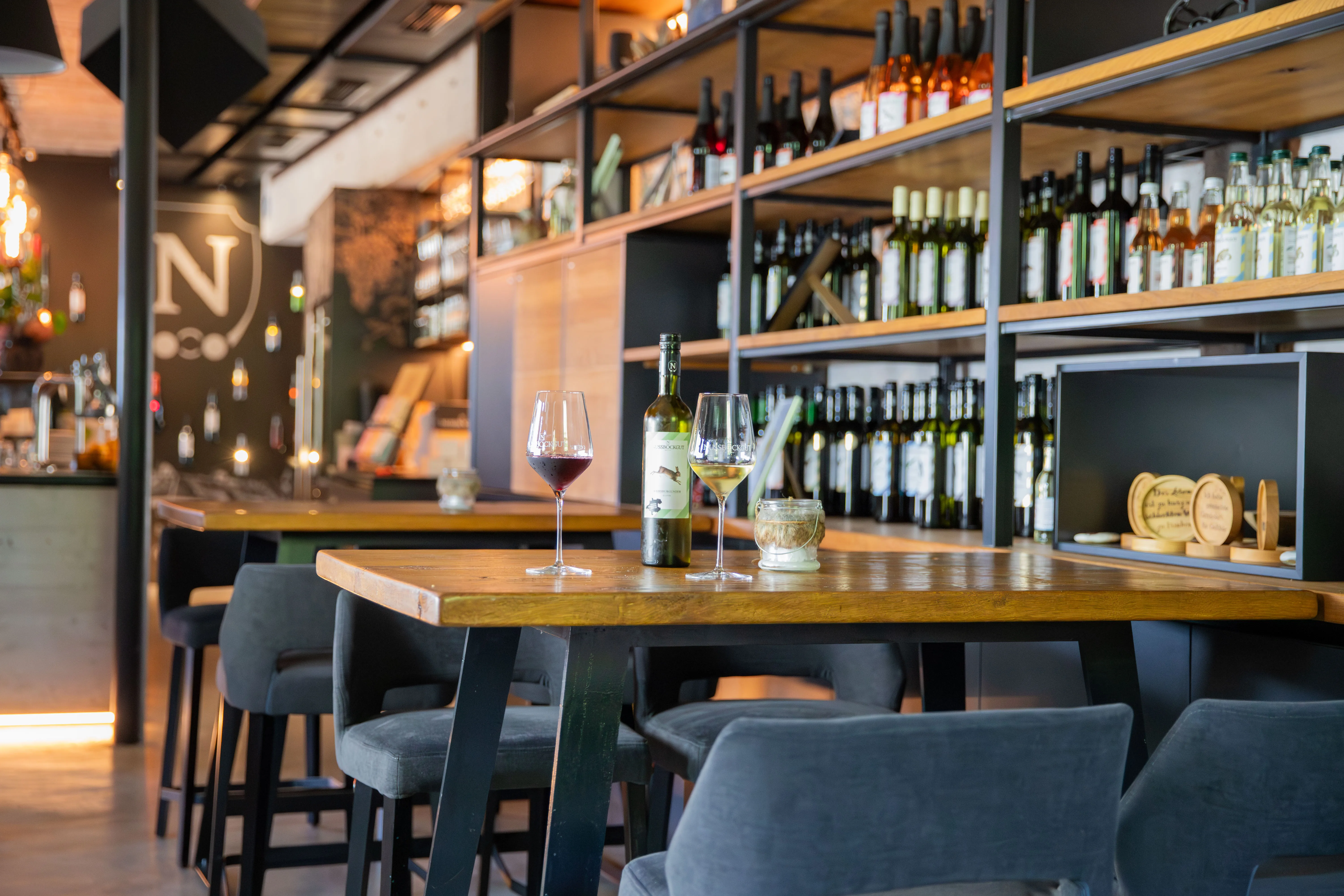 Ein brauner Tisch mit Weingläsern und einer Flasche, grauen Sesseln; im Hintergrund Regale mit Flaschen und weiterer Tisch bzw. Theke