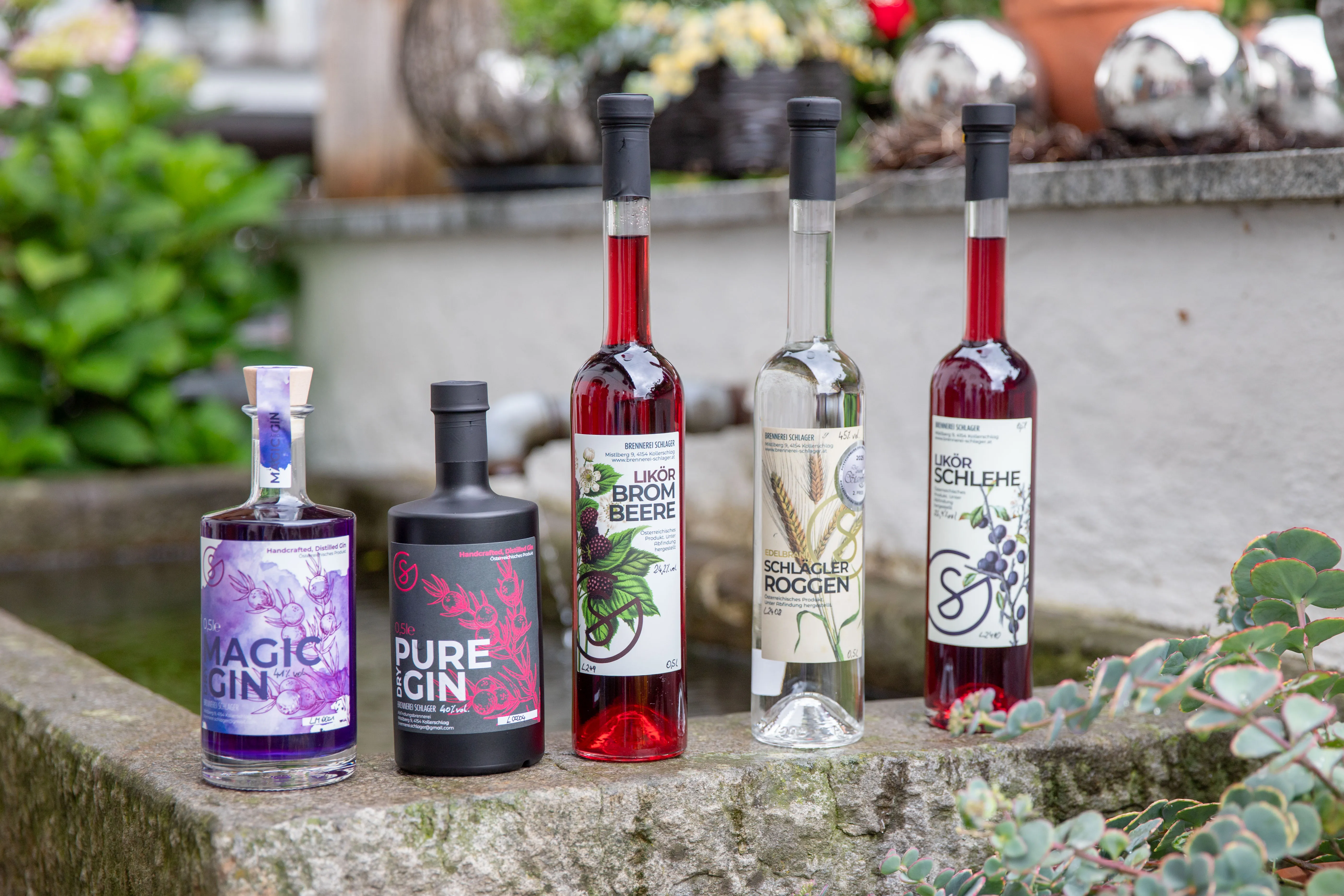 Fünf verschieden färbige Flaschen stehen auf einem grauen Steinrand eines Steingranders, im Hintergrund Dekor und grüne Büsche
