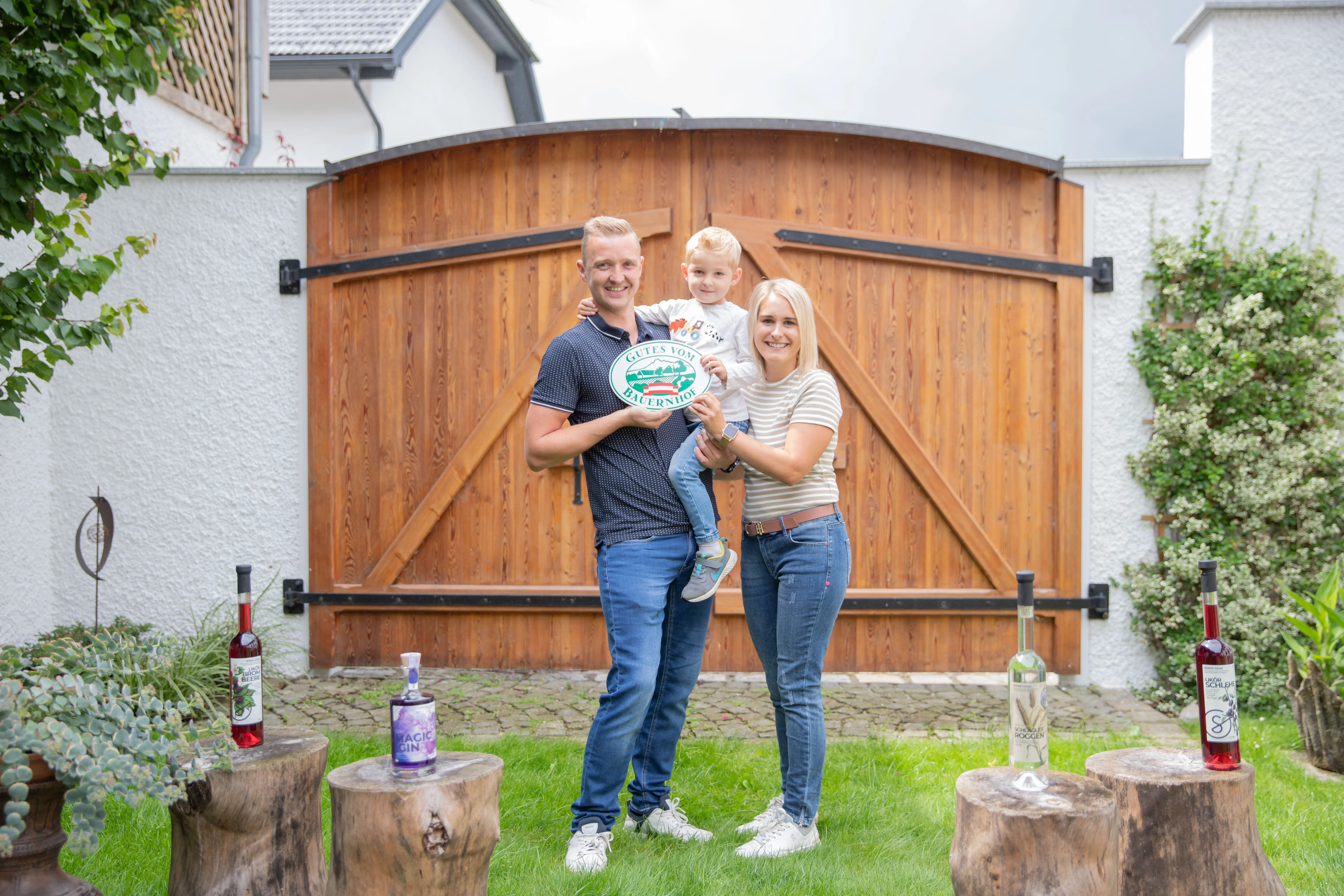 Vater, Mutter und Kind auf dem Arm stehen vor dem braunen Hoftor auf der grünen Wiese; im Vordergrund Baumstümpfe mit einzelnen Flaschen darauf
