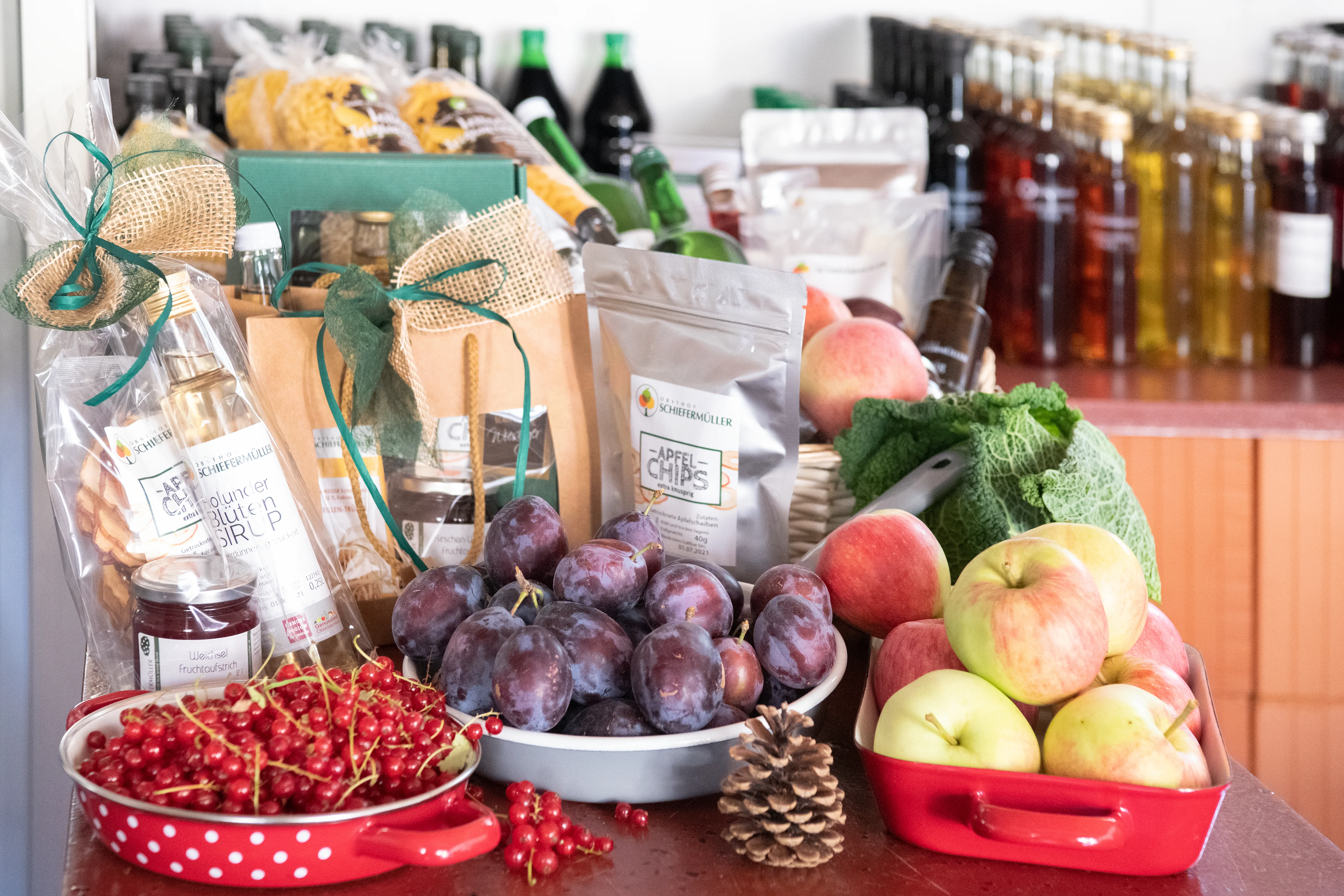 Auf eine Tisch stehen Produkte und Schalen mit Ribisel, Zwetschken und Äpfel, dahinter eine Theke mit Flaschen und weiteren Produkten,  weiße Wand