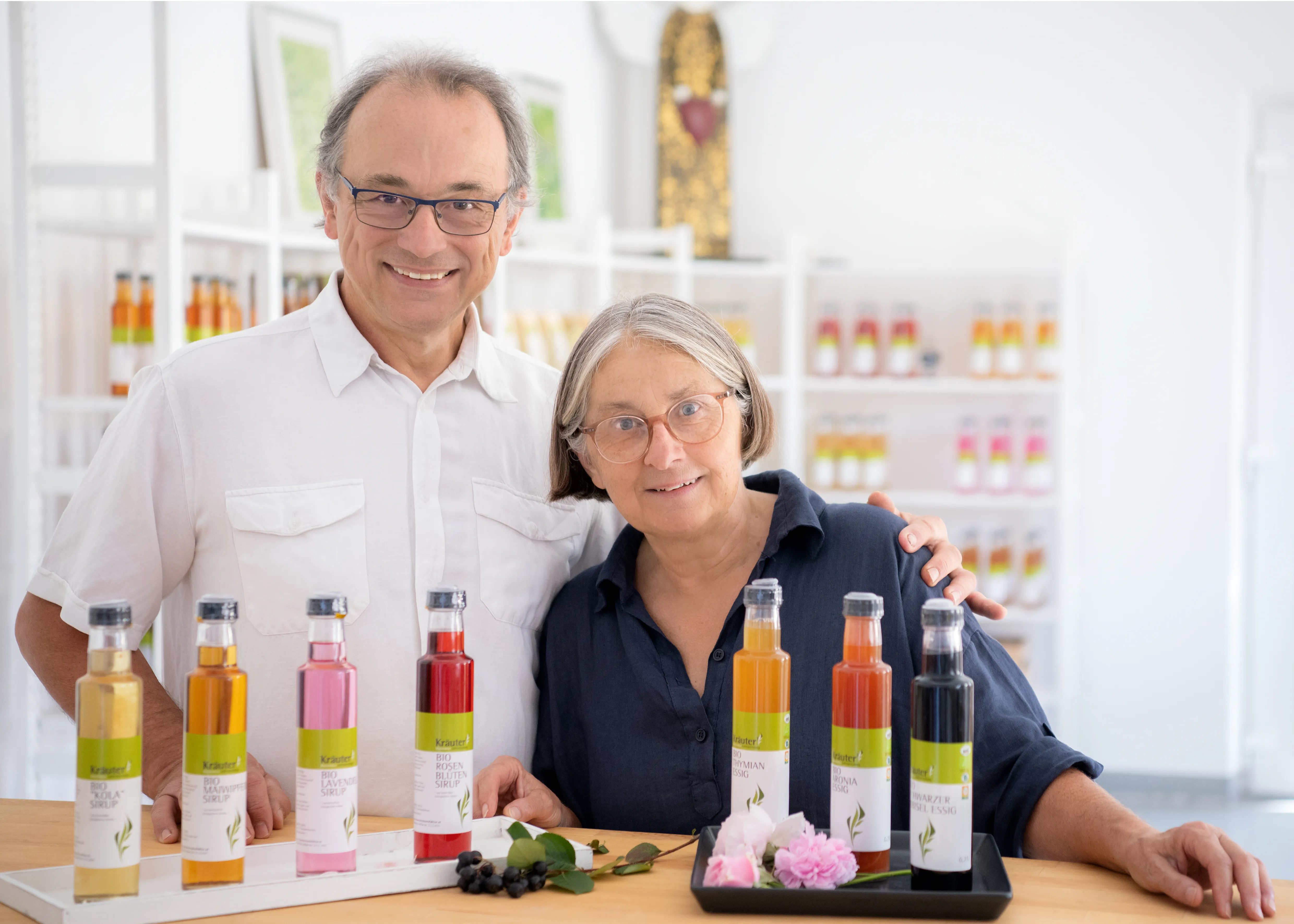 Ein Mann und eine Frau stehen hinter einer Theke mit verschiedenen Flaschen, daneben Kräuter, dahinter weiße Regale mit weiteren Flaschen