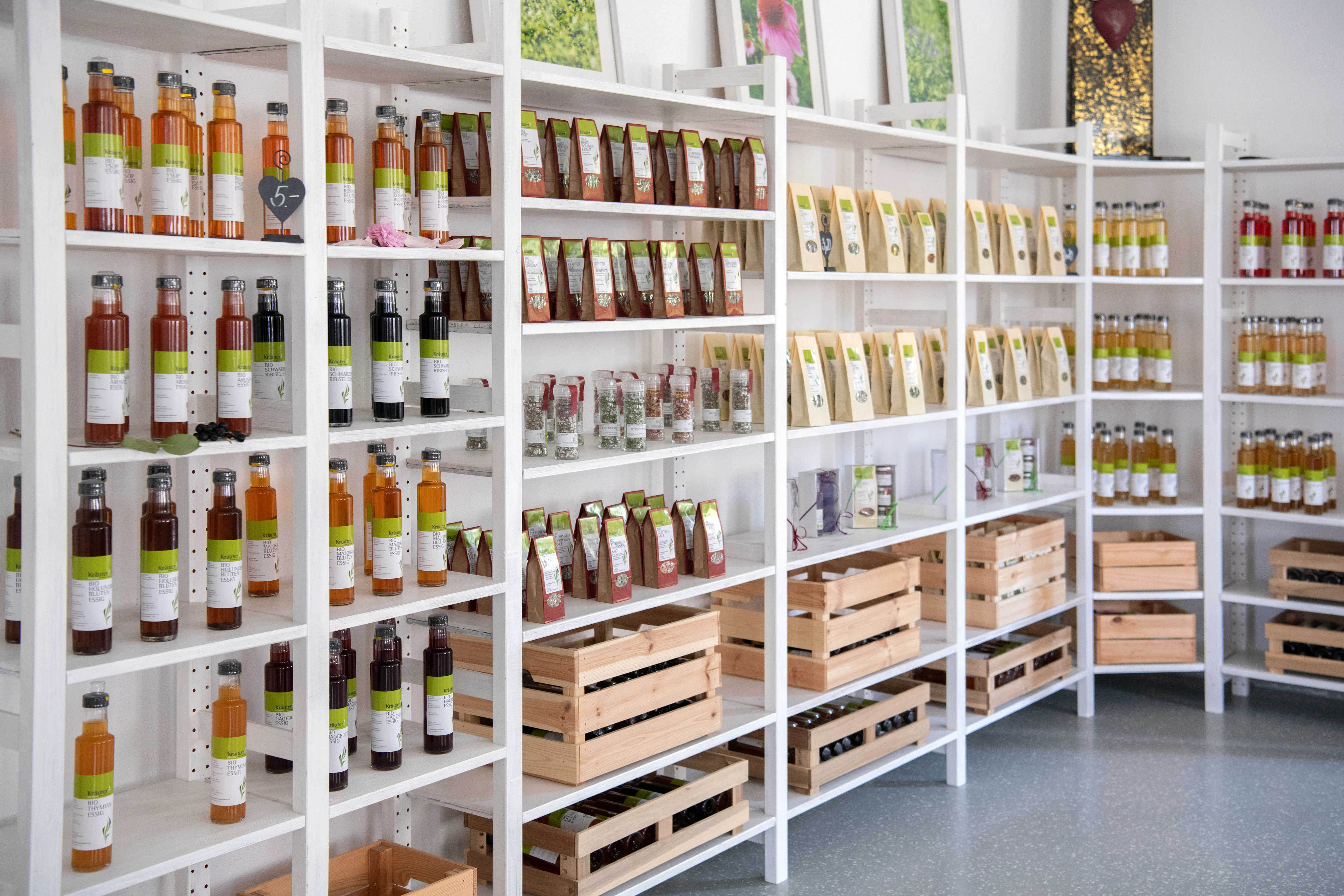 An einer Wand stehen weiße Regale mit diversen Flaschen und Verpackungen, Holzkisten, ganz oben Bilder von Kräuterfeldern