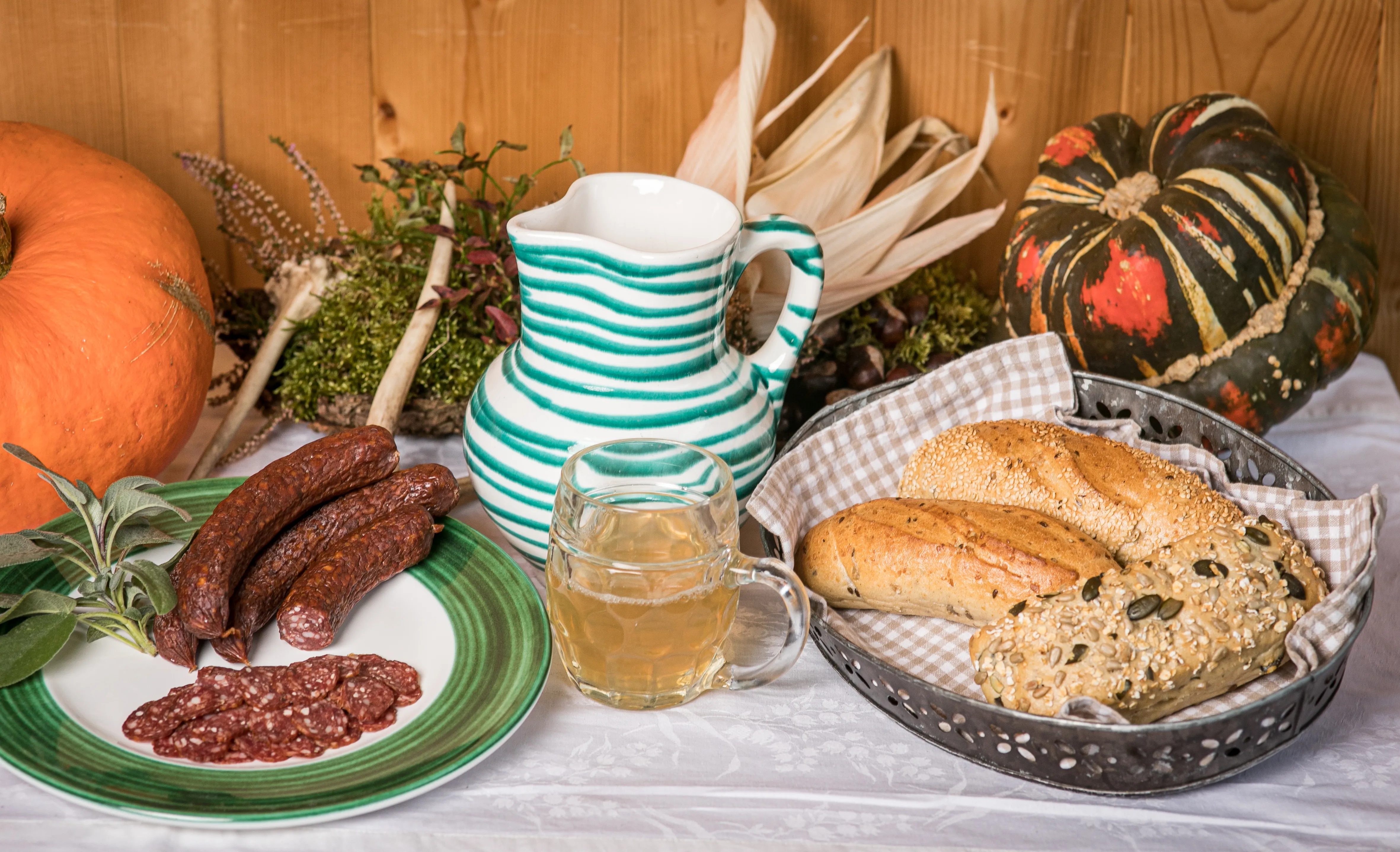 Auf einem Tisch stehen ein Krug, ein Glas und zwei Teller mit Würsten und Gebäck; daneben liegen zwei Kürbisse, ein Maiskolben und Kräuter