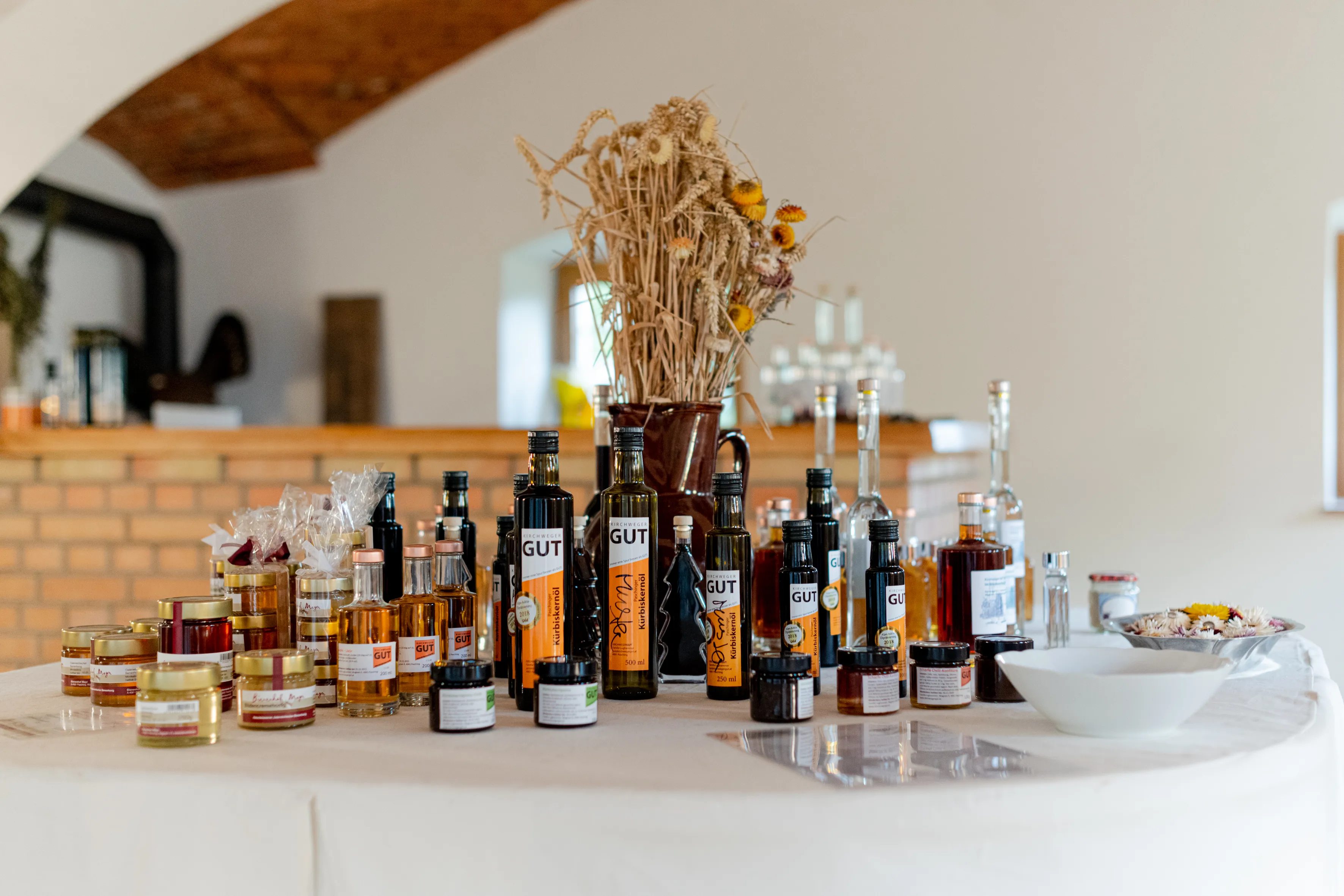 Verschieden Flaschen, Gläser und Produkte stehen auf einem Tisch mit weißem Tischtuch, dahinter Ziegeltheke, getrocknete Gräser in einer Vase