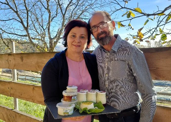 Ein Mann mit rosa Shirt und dunkler Weste und ein Mann im grauen Hemd mit Brille stehen vor einem Holzzaun und halten ein Tablett mit Milchprodukten in der Hand. Dahinter grüne Wiese und kahle Bäume