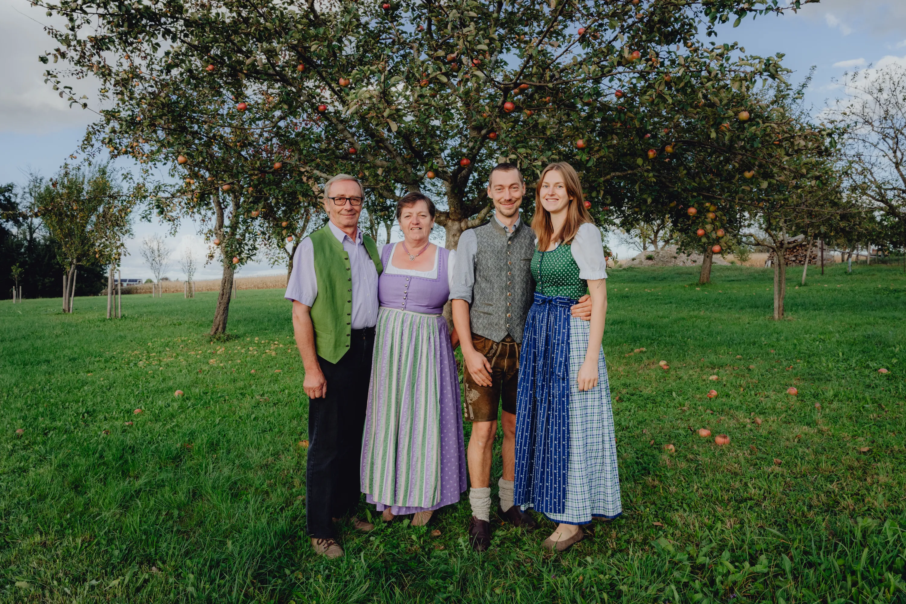 Zwei Frauen und zwei Männer, gekleidet in Tracht, stehen auf einer grünen Wiese, im Hintergrund Bäume