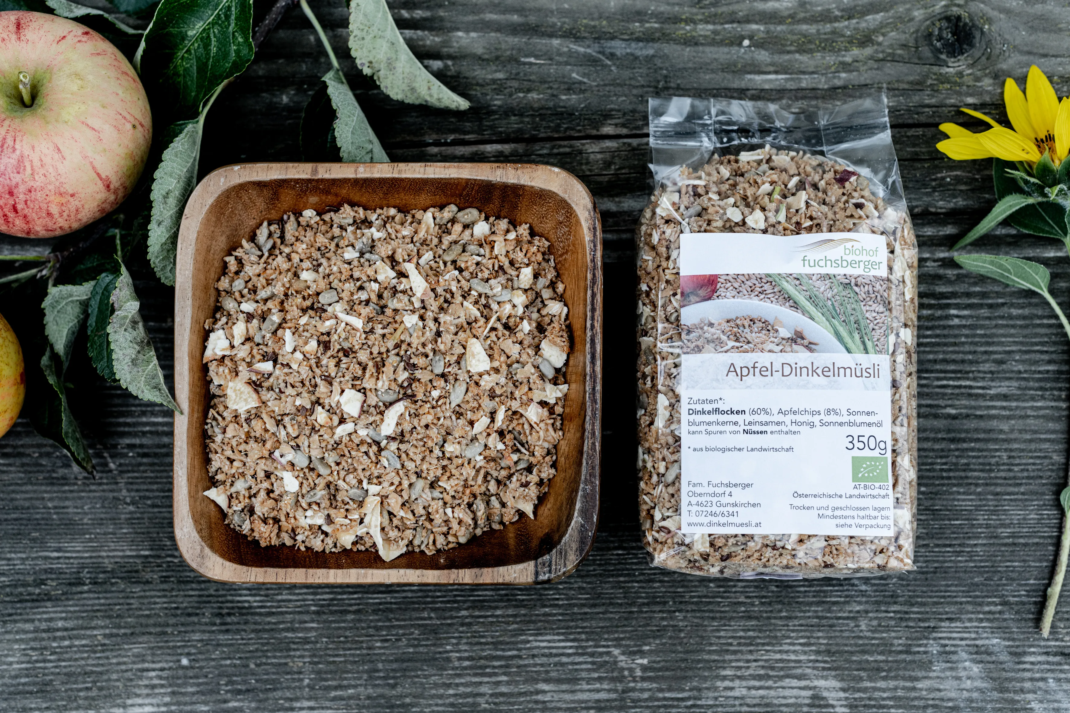Auf einem schwarzen Hintergrund sind eine Schale und ein Sackerl mit Müsli zu sehen, daneben  ein Apfel und eine Sonnenblume