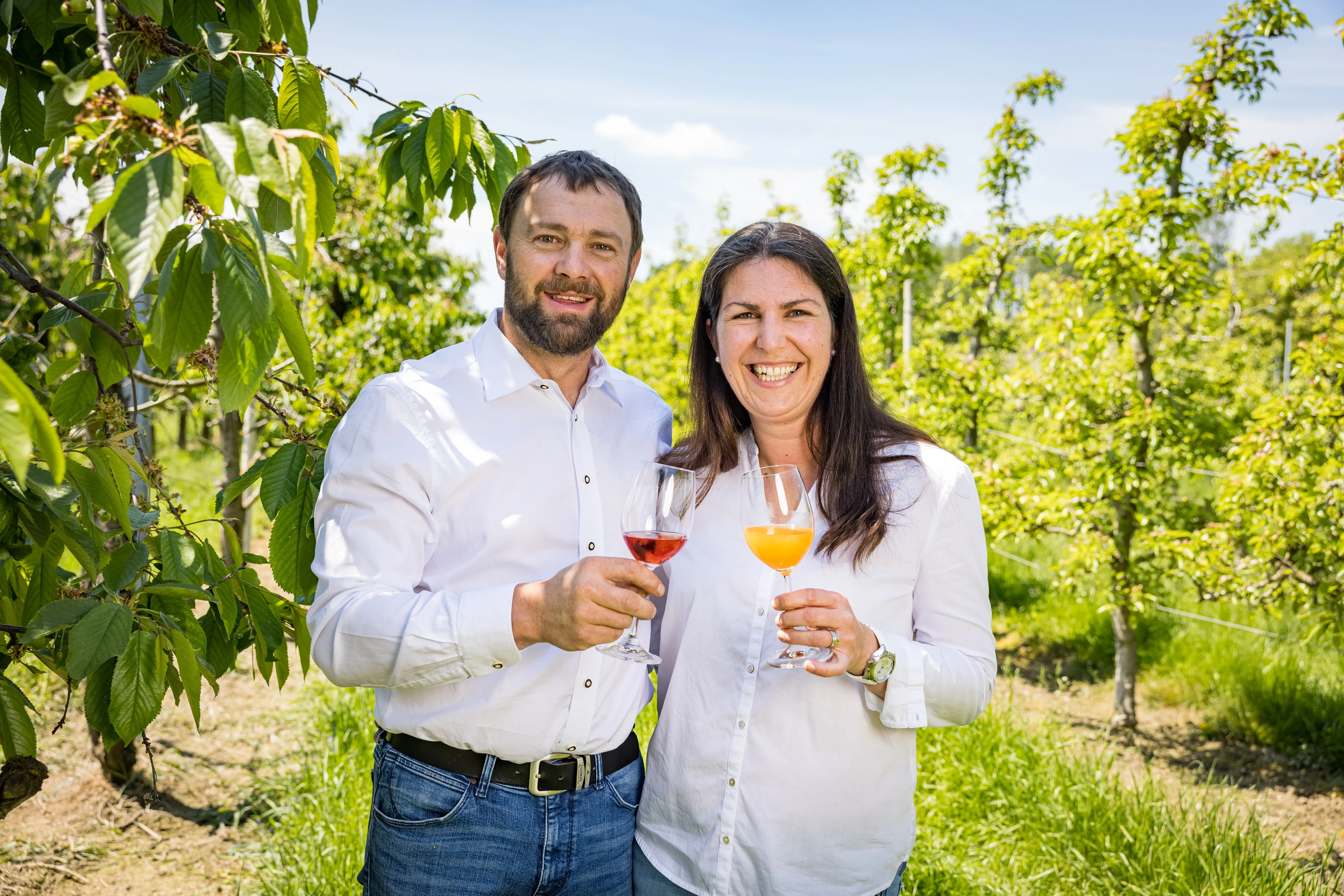 Ein Mann und eine Frau in weißem Hemd bzw. Bluse und Jeans stehen in einer Obstplantage und halten ein Glas mit rotem und orangem Inhalt in der Hand. Dahinter Obstbäume und grüne Wiese