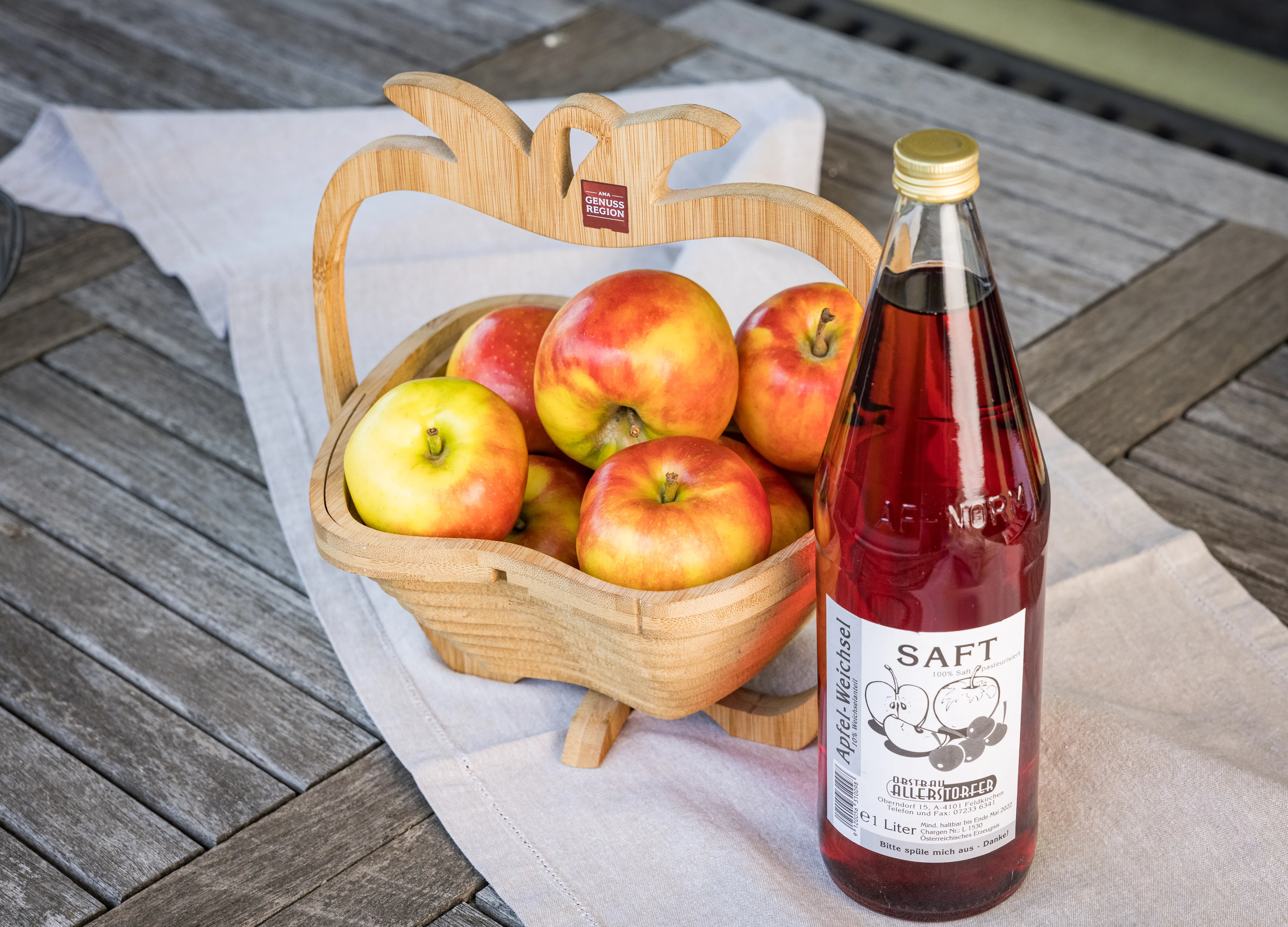 Auf einem Holztisch liegt eine weiße Decke, darauf ein steht Holzkorb mit gelb/roten Äpfeln, daneben eine Flasche mit rotem Inhalt 