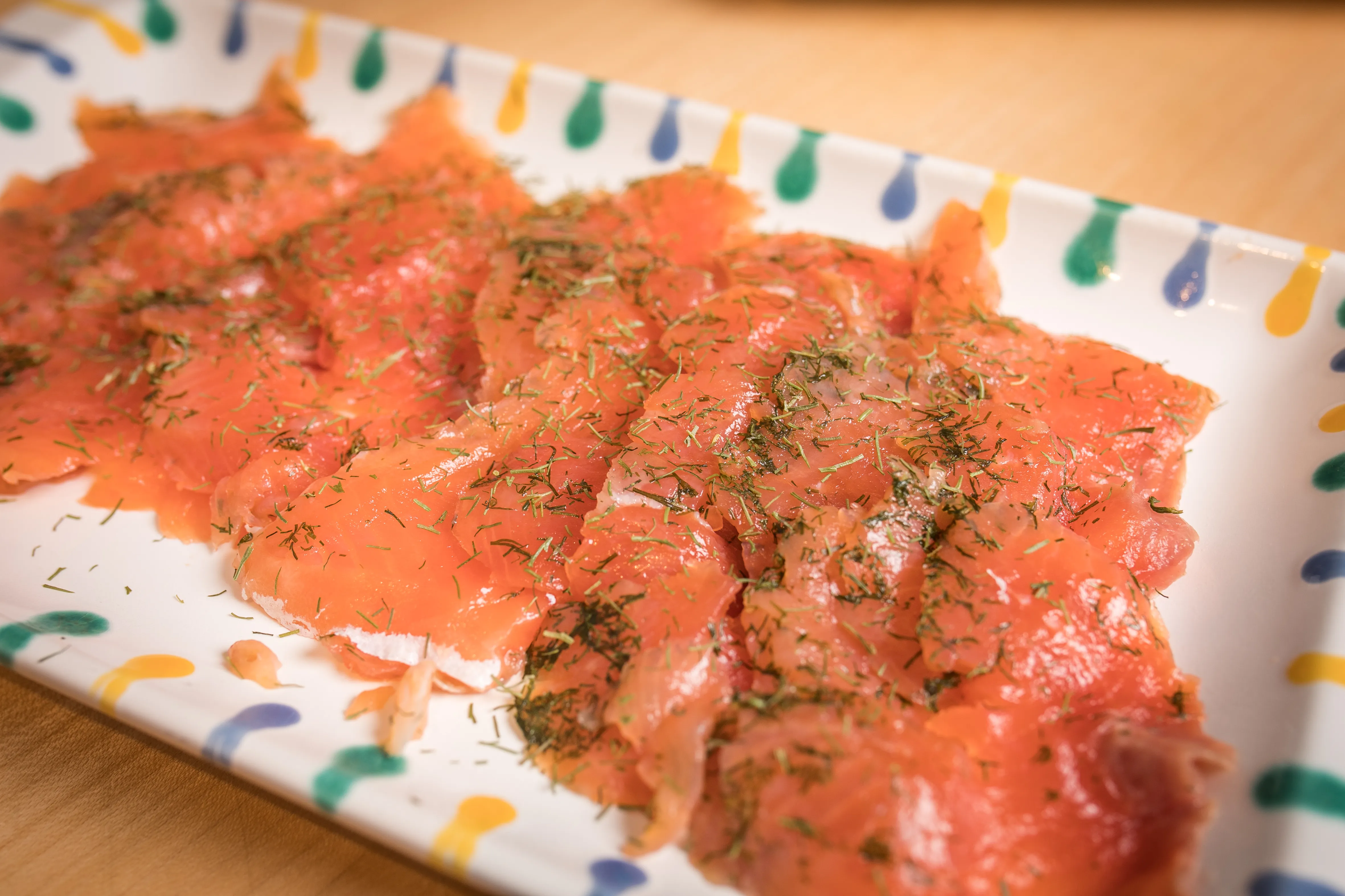 Auf einer weißen Schale mit gelben, grünen und blauen Strichen liegt orangefarbener Lachs mit Dill