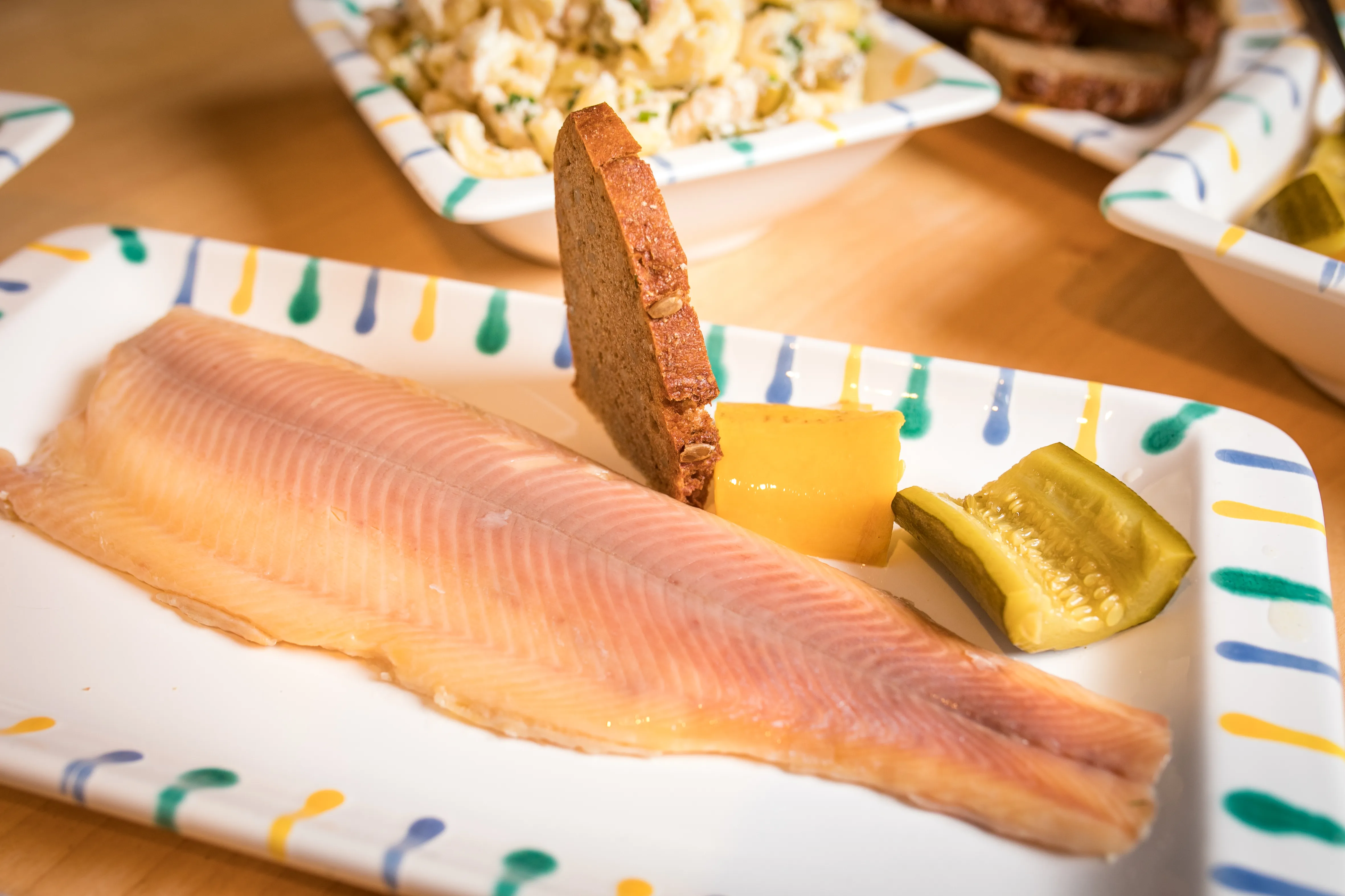 Auf einer weißen Platte liegt ein Fischfilet, garniert it Gurke und Brot, dahinter reine Schüssel Kartoffelsalat