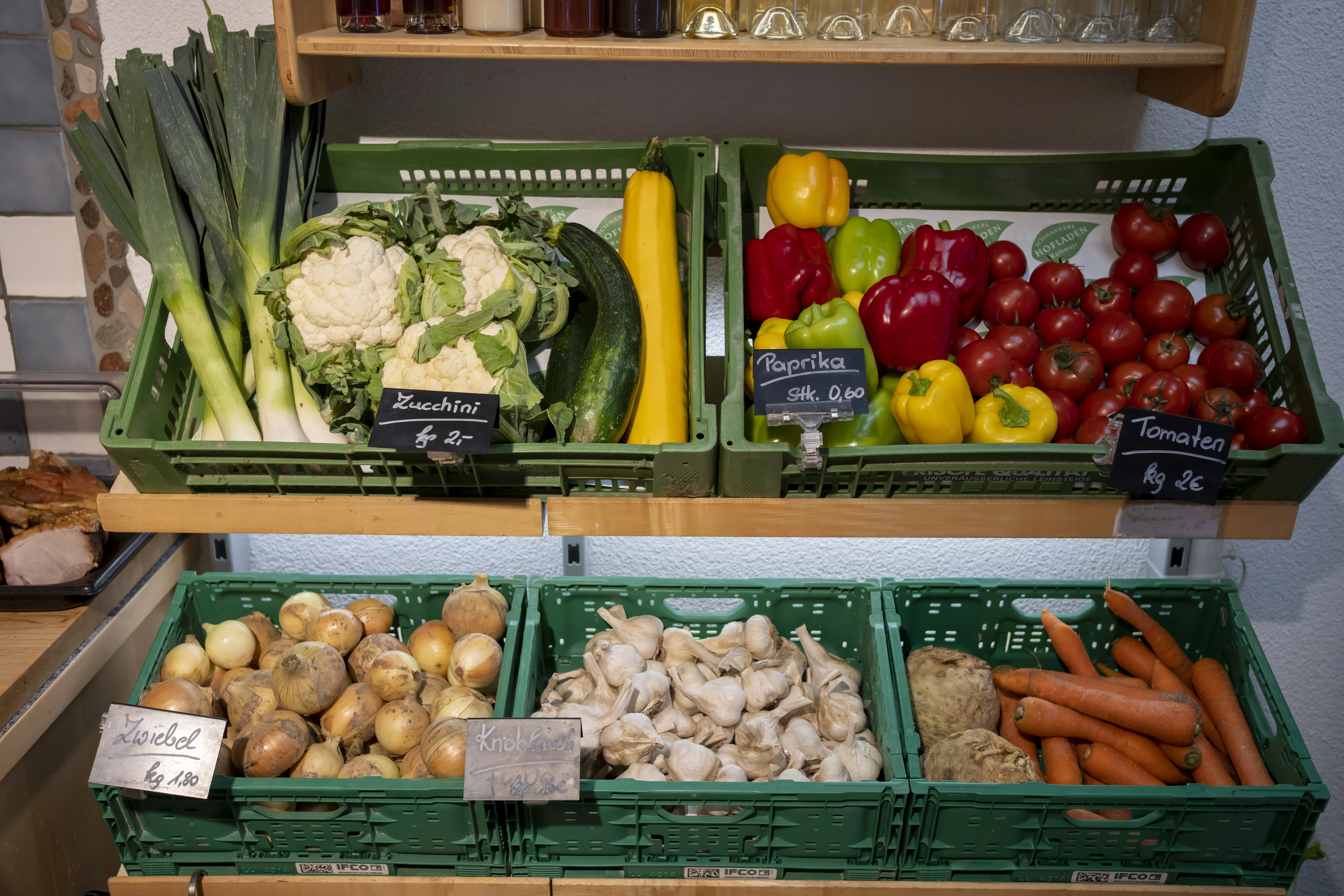 Auf einem braunen Regal stehen grüne Kisten mit verschiedenem Gemüse drin (Karotten, Sellerie, Knoblauch, Zwiebel, Lauch, Blumenkohl, Paprika, Zucchini)