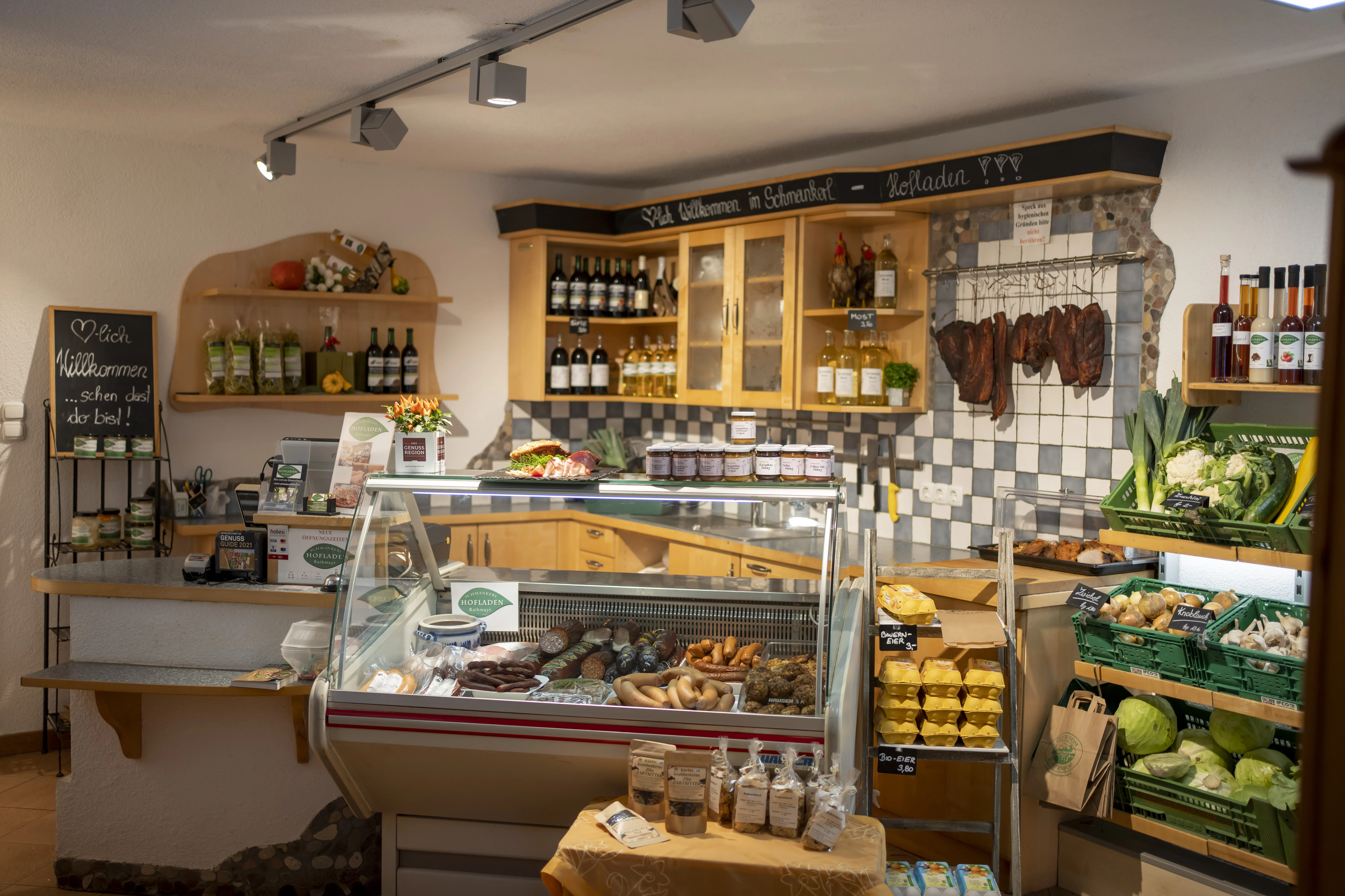 Mehrere Regale, Hängekästen an der Wand, davor eine Kühlvitrine mit verschiedenen Produkten (Gläser, Flaschen, Fleischprodukte, Gemüse, Eier)