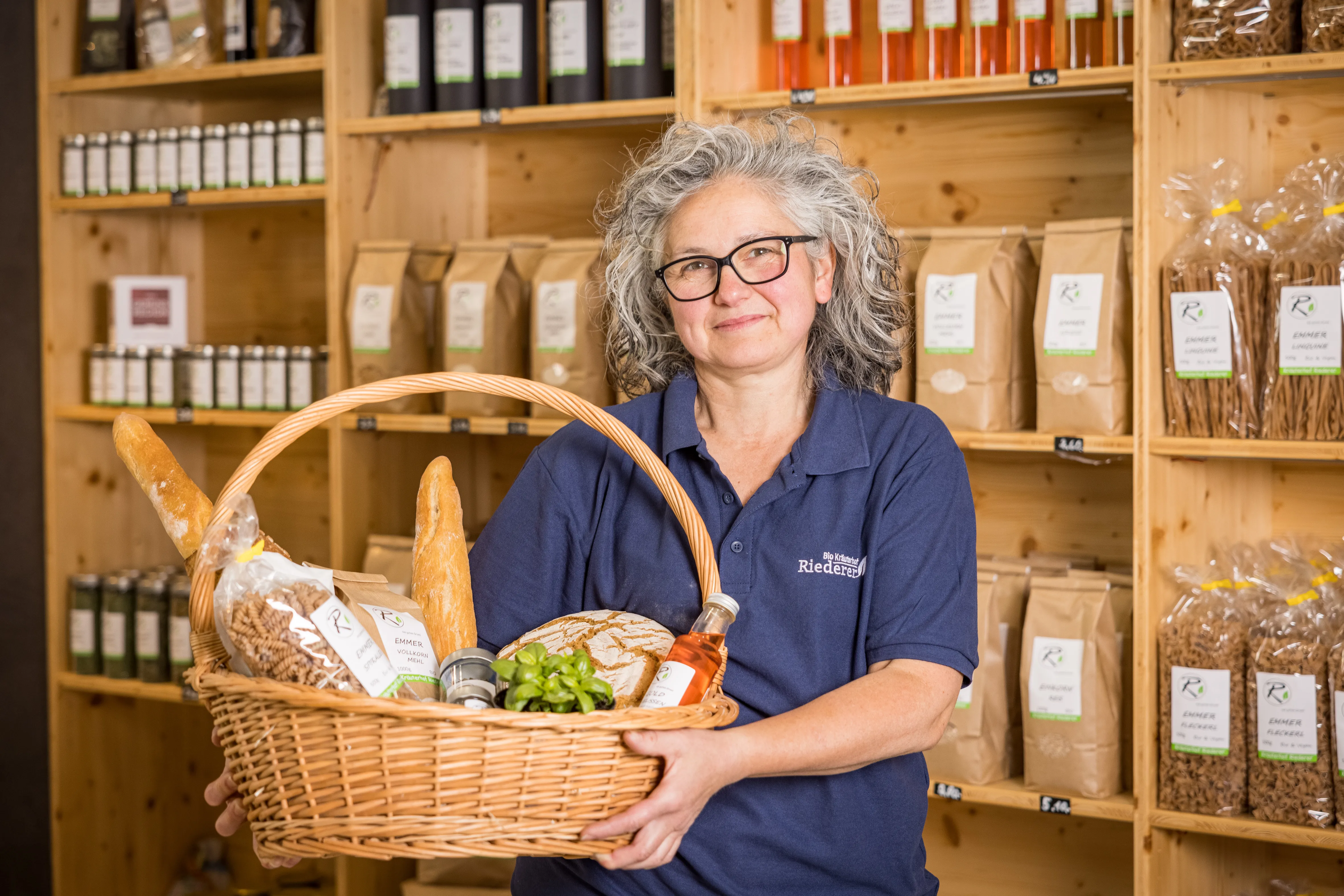 Eine grauhaarige Frau mit blauem Polo-Shirt hält einen Korb in den Händen. Im Korb befinden sich Baguette, Nudeln, Brot eine Flasche, ein Glas, ein Sackerl und Kräuter. Im Hintergrund ein Holzregal an der Wand mit verschiedenen Flaschen, Gläsern und Papiersackerl.