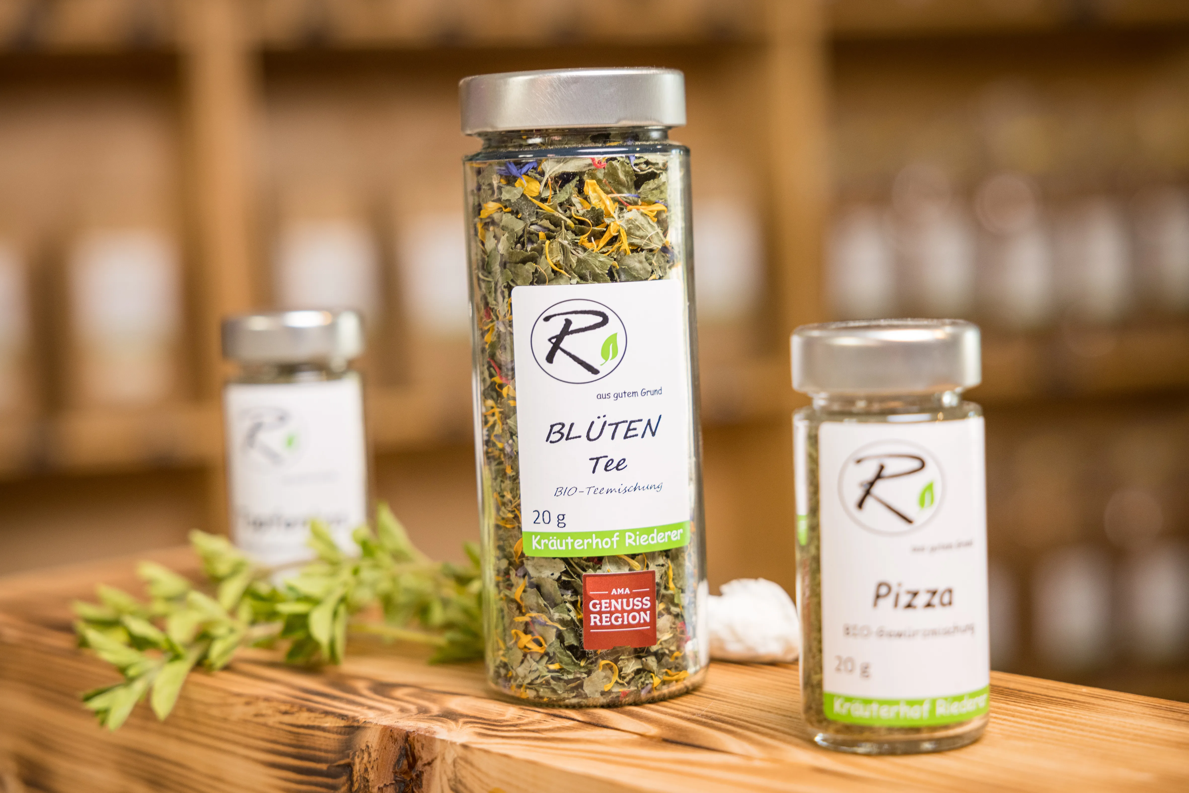 Auf einem Holztisch stehen drei Gläser mit Blüten und Kräutern, daneben frische Kräuter. Im Hintergrund etwas verschwommen ein Regal.