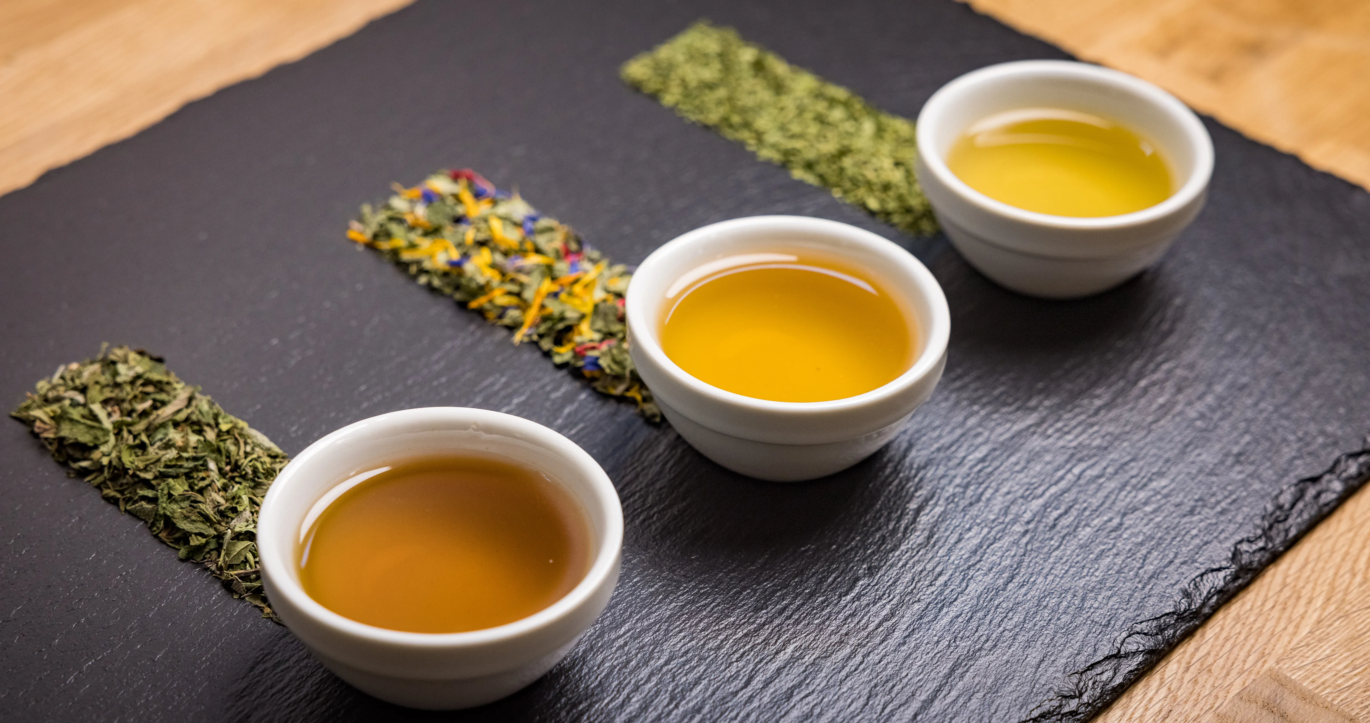 Auf einer schwarzen Platte auf einem Holztisch liegen bzw. stehen drei weiße Schalen mit gelber bzw. orangefarbener Flüssigkeit, daneben frische Kräuter.