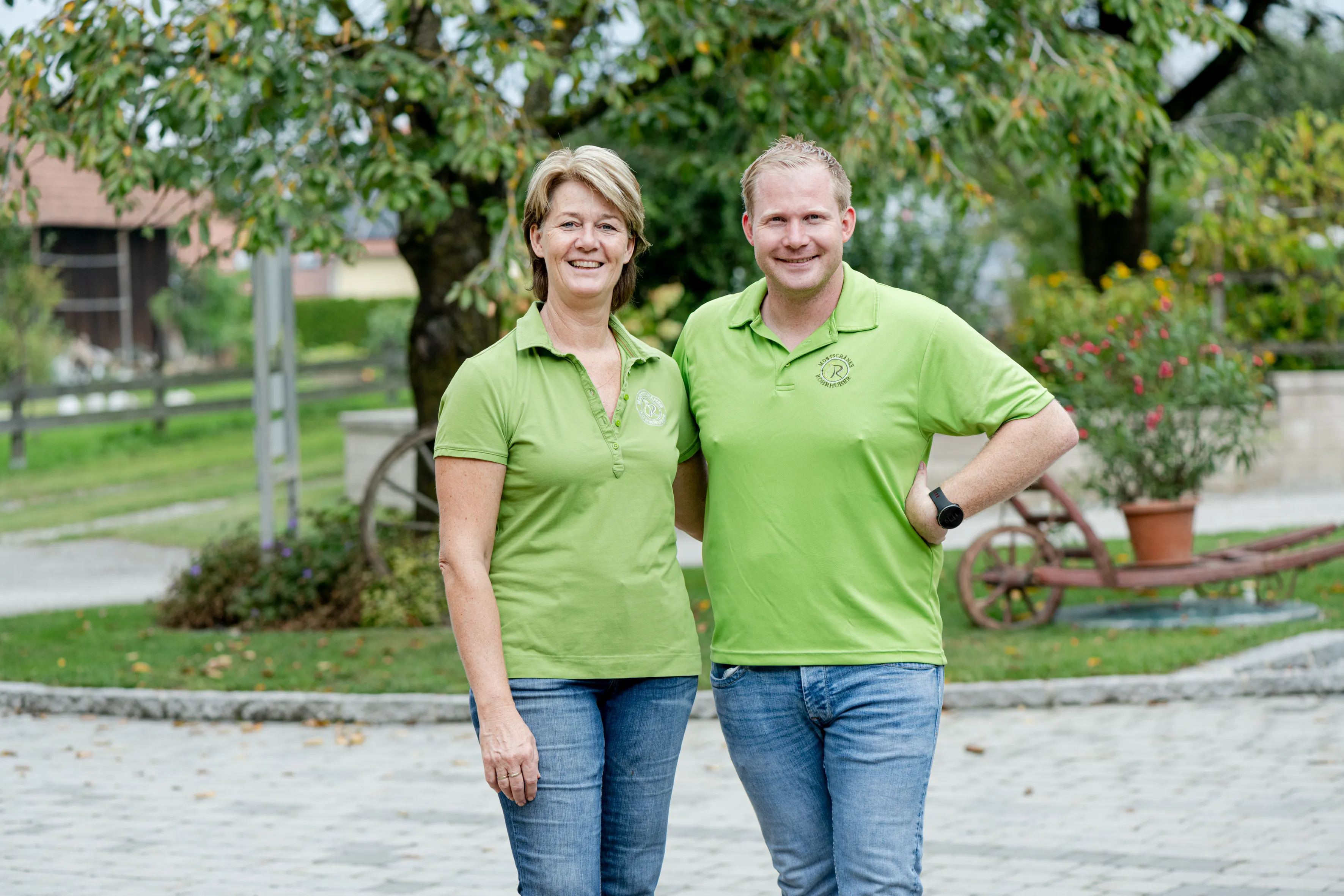 Ein Mann und eine Frau in grünem T-Shirt und Jeans stehen vor einem Baum und Wiese auf Pflastersteinen