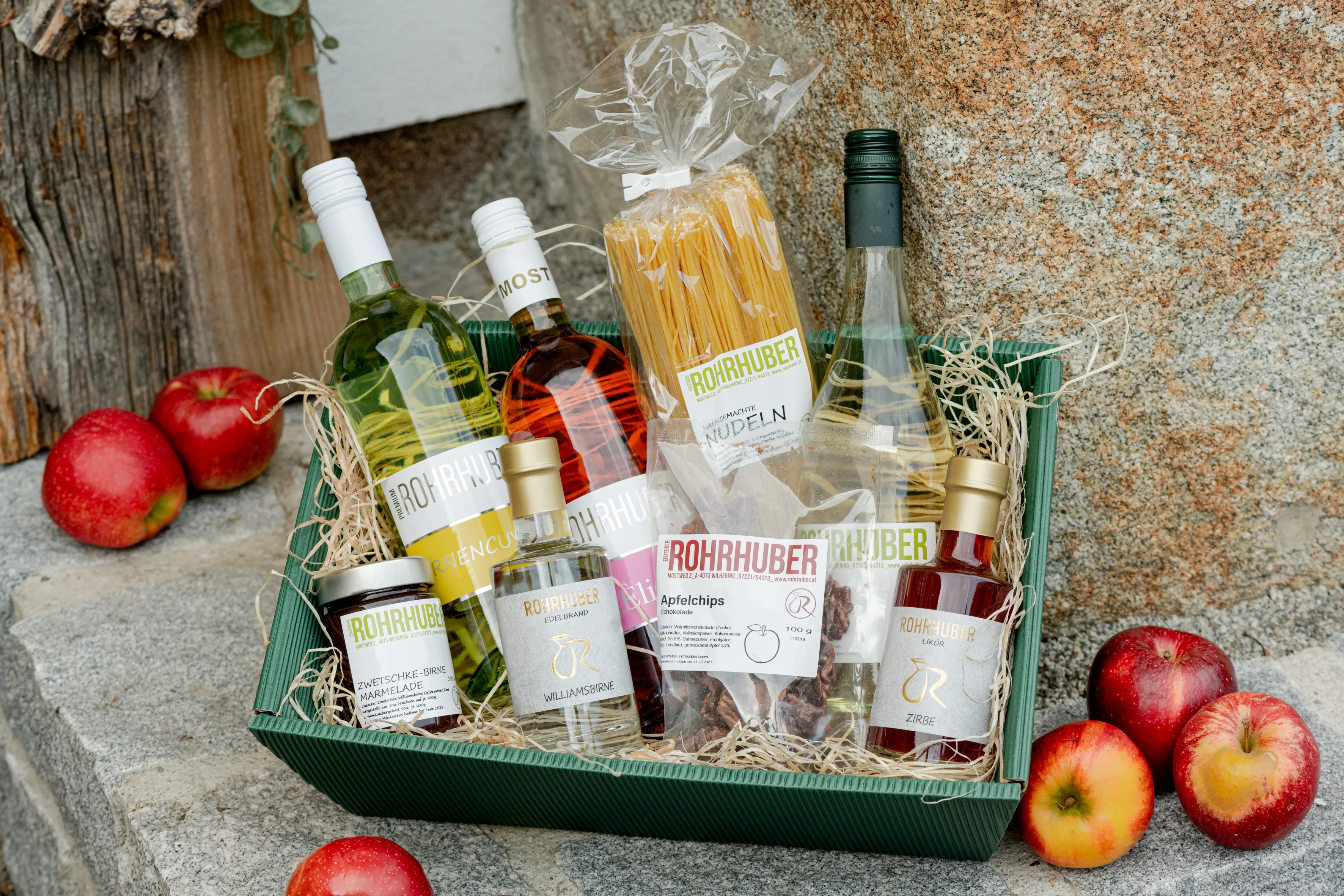 Ein Geschenkkorb mit Flaschen und Stroh steht auf einer Steinplatte, daneben liegen Äpfel