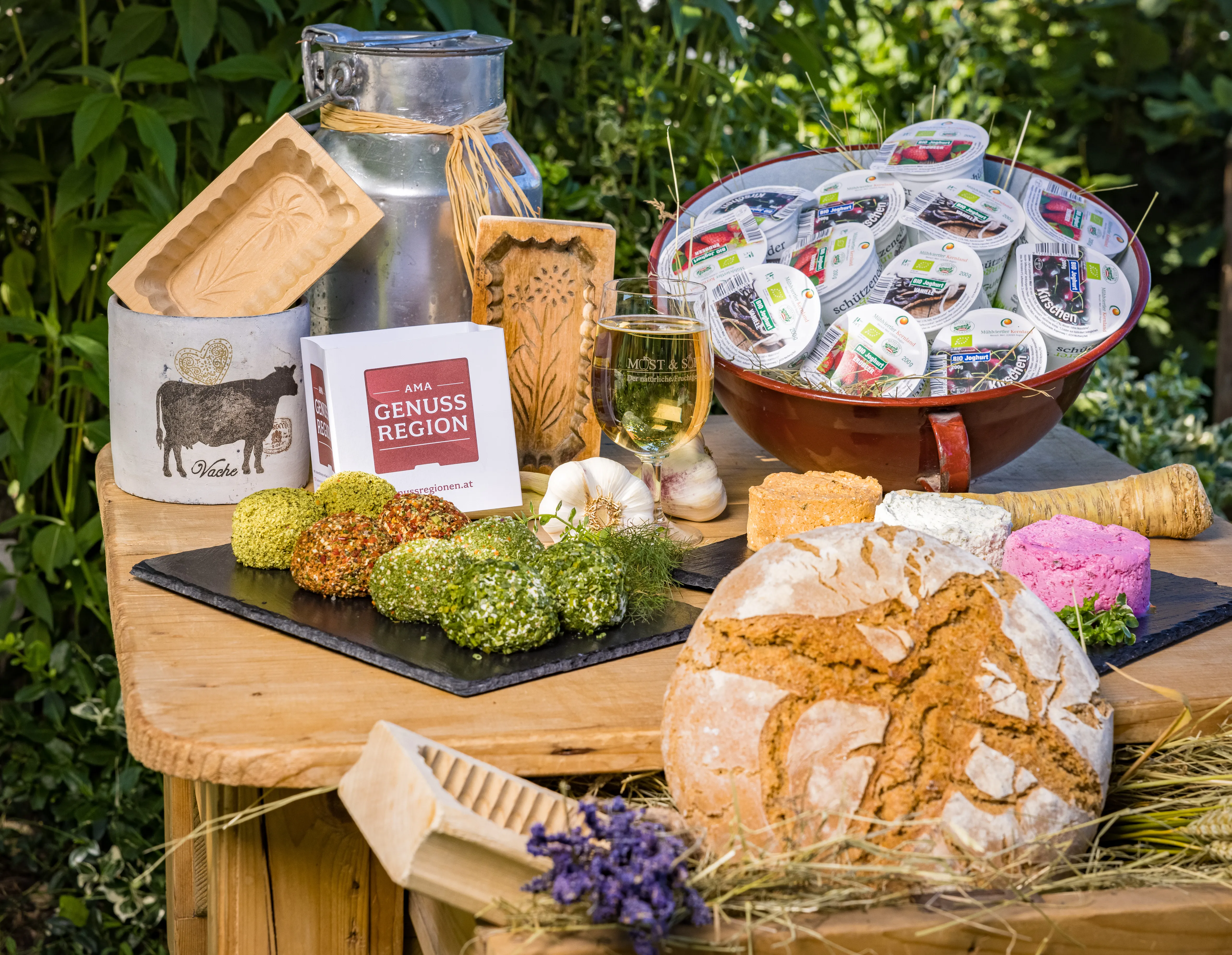 Auf einem Holztisch liegen bzw. stehen eine Milchkanne, ein Häferl, eine Platte mit Frischkäse, eine roter Weidling mit Joghurt, ein Laib Brot, dazwischen liegen Kräuter, Stroh und Knoblauch. Ebenfalls dazwischen ein Mostglas mit Inhalt. Dahinter grünes Blattwerk.